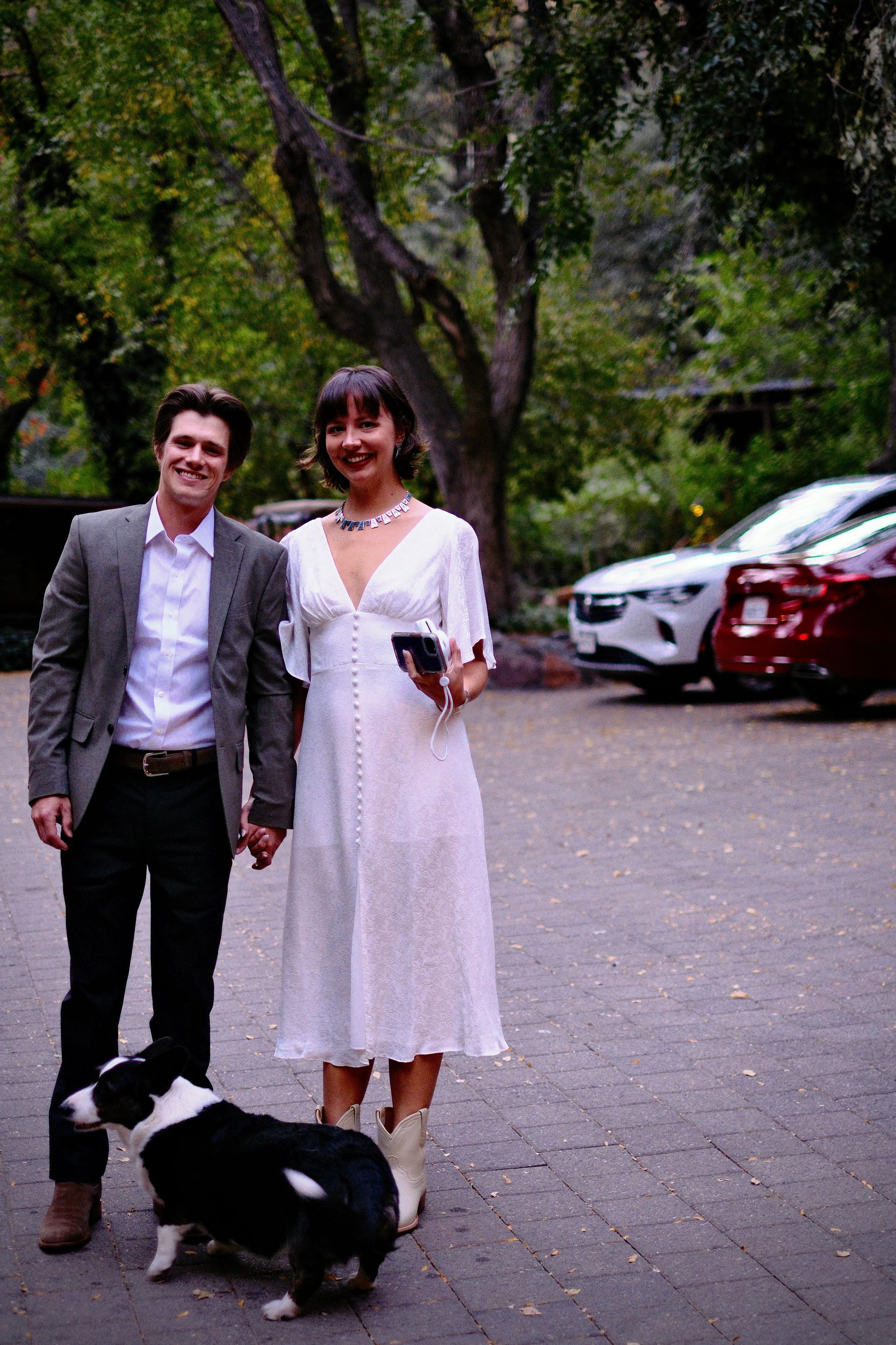 Two people standing outdoors with a black-and-white dog, dressed formally near parked cars and trees.