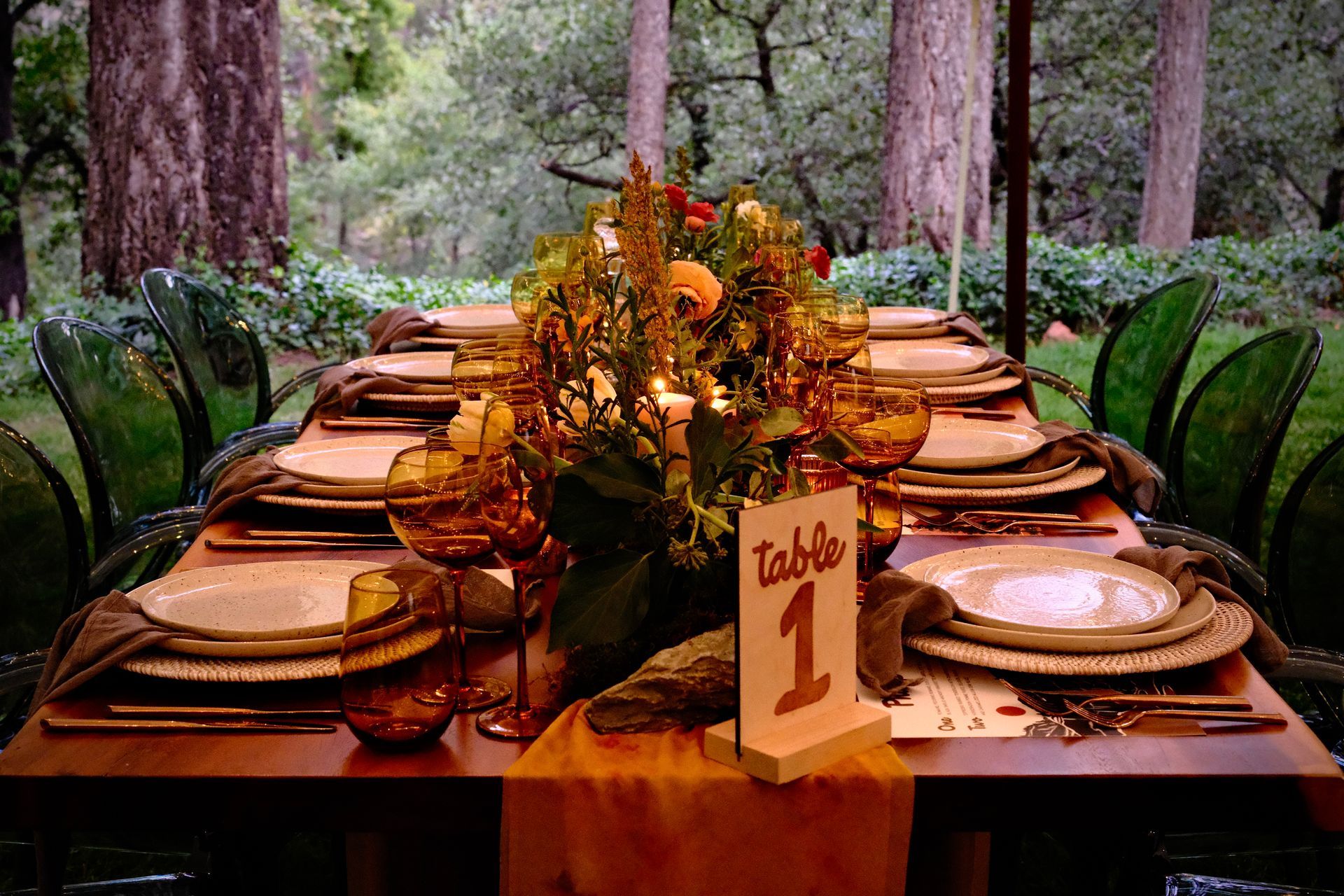 Elegant outdoor banquet table set in a forest, with candles, flowers, and a table number sign.