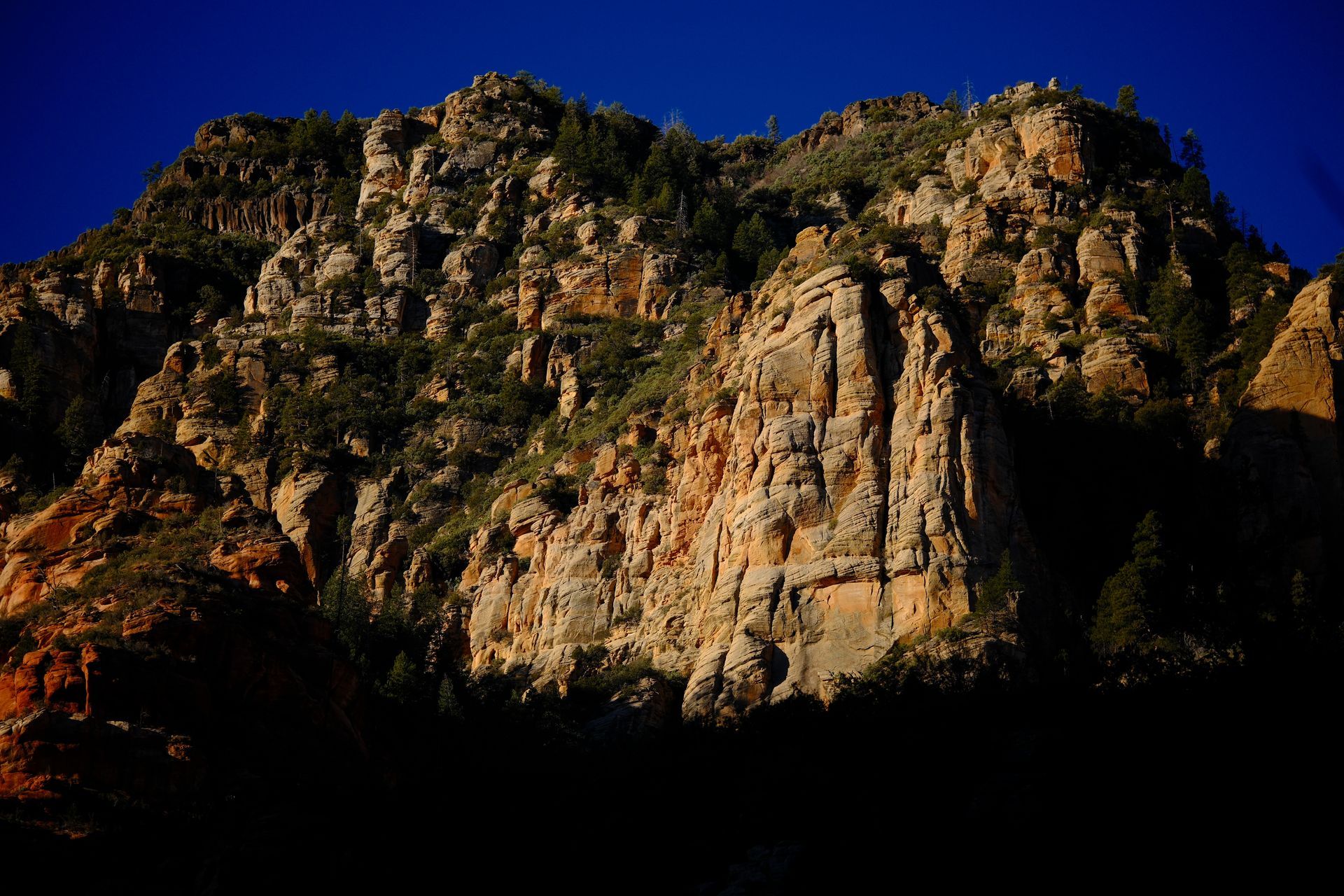 Sunlit rocky cliff face against a deep blue sky, with dark shadowed foreground