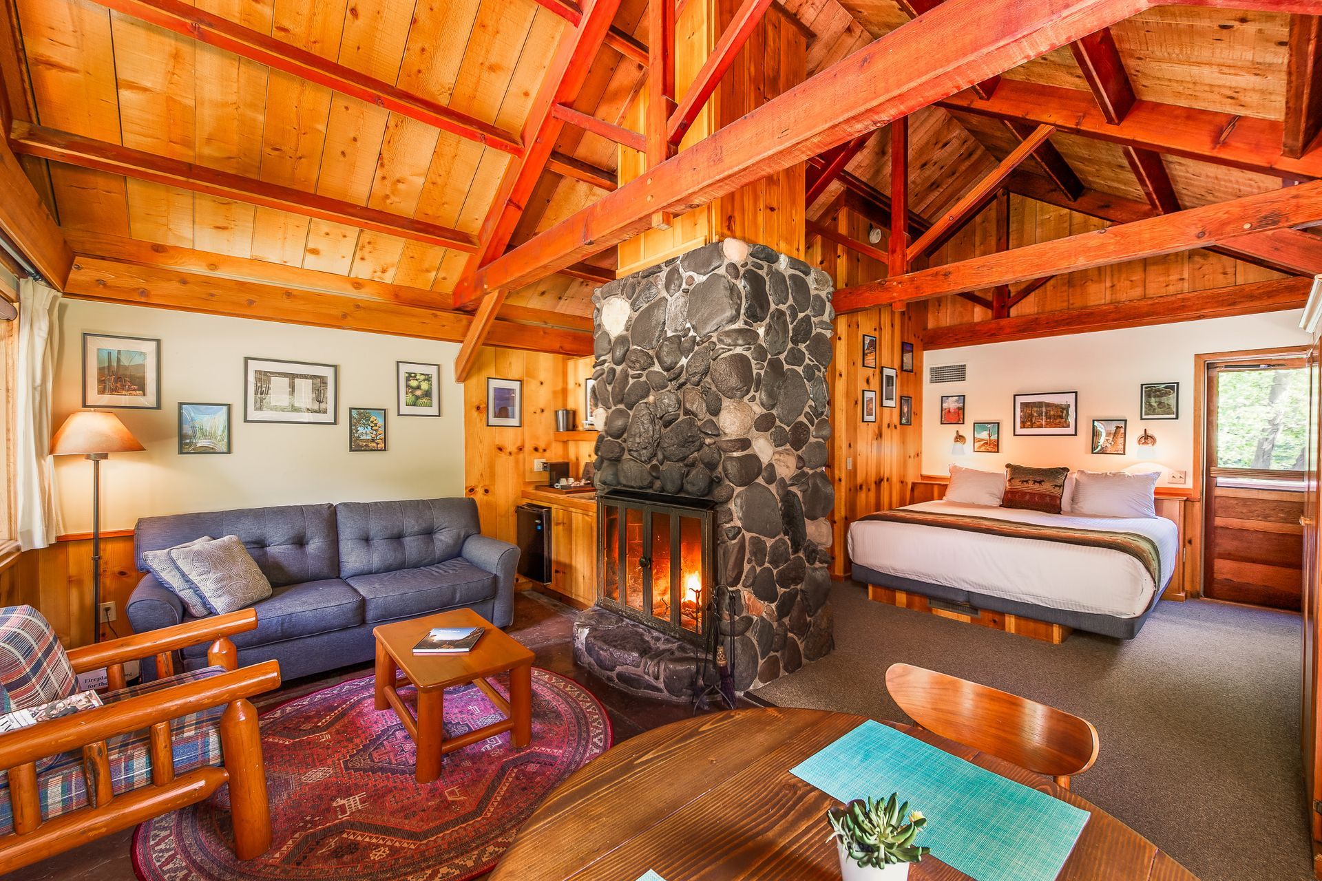 Rustic lodge living room with wood beams, stone fireplace, blue sofa, and bed by windows