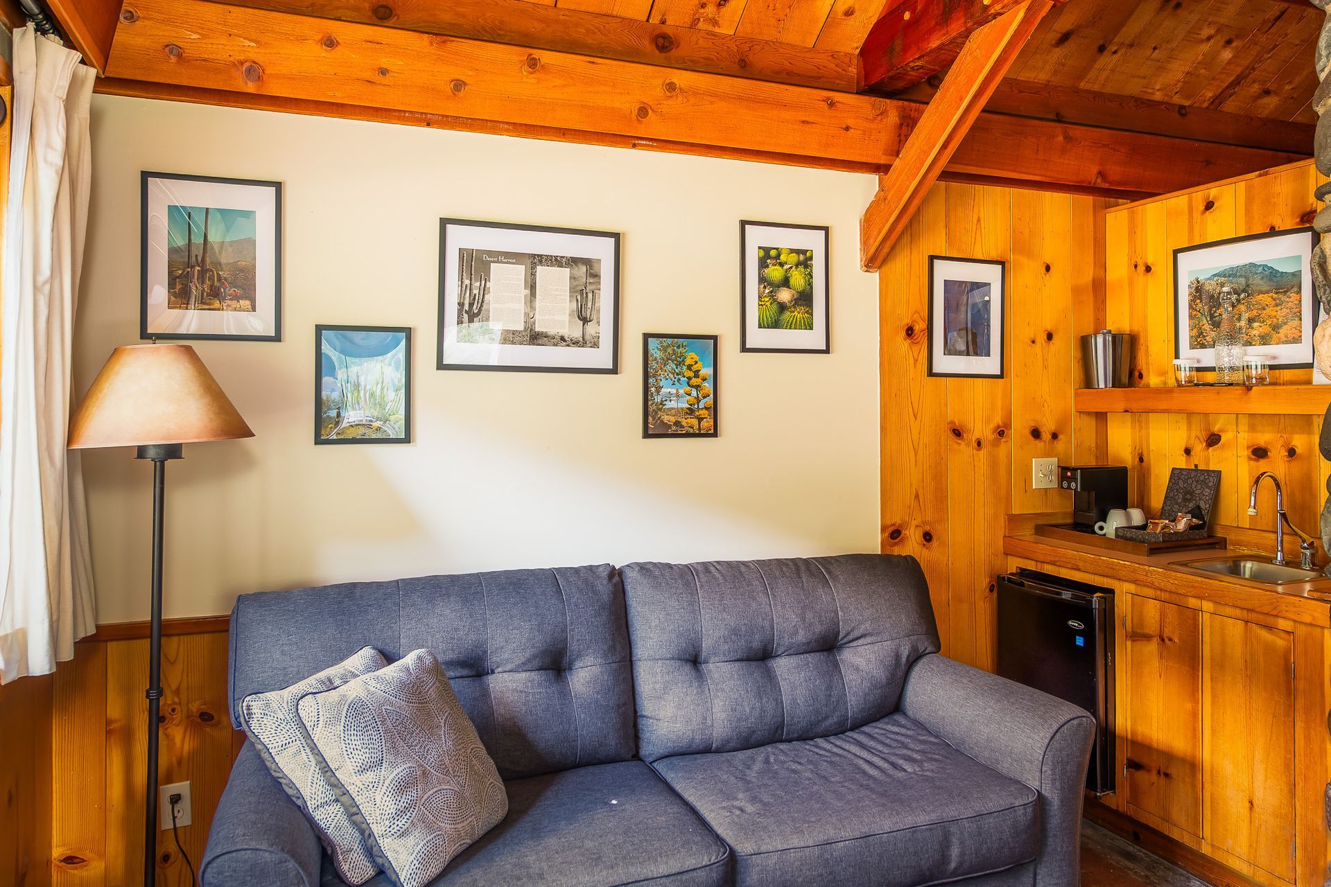 Cozy cabin living room with blue sofa, wood walls, framed art, and a small kitchen nook.