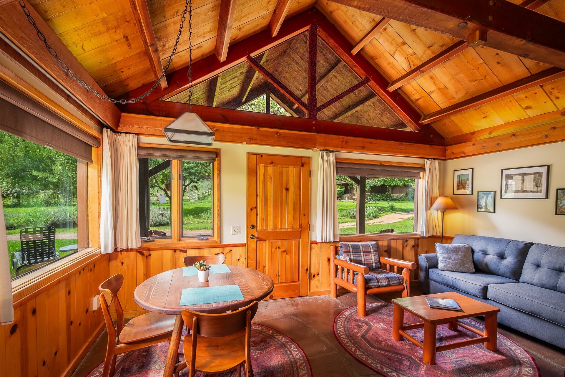 Rustic cabin living room with wood beams, large windows, dining table, and gray sofa