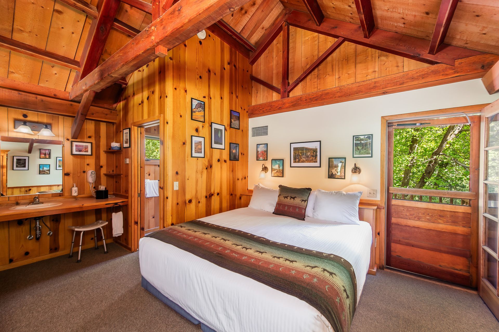 Wood-paneled cabin bedroom with a white bed, wooden ceiling beams, and a doorway to greenery.