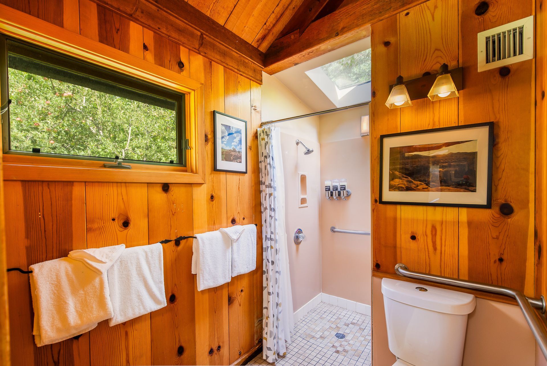 Wood-paneled rustic bathroom with towels, toilet, shower, and small window looking onto greenery