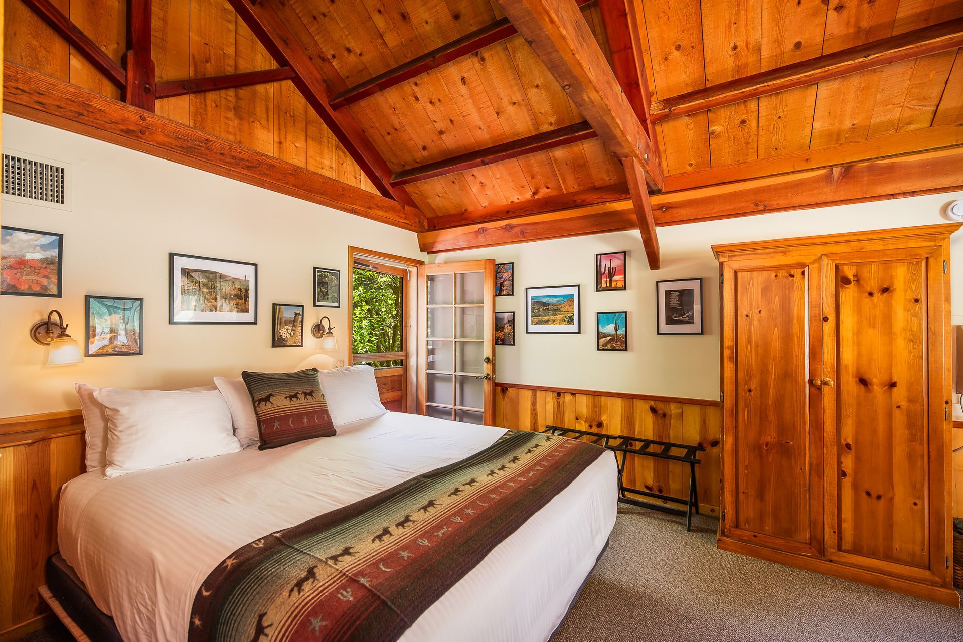 Cozy wood-paneled bedroom with vaulted ceiling, queen bed, framed wall art, and wooden wardrobe