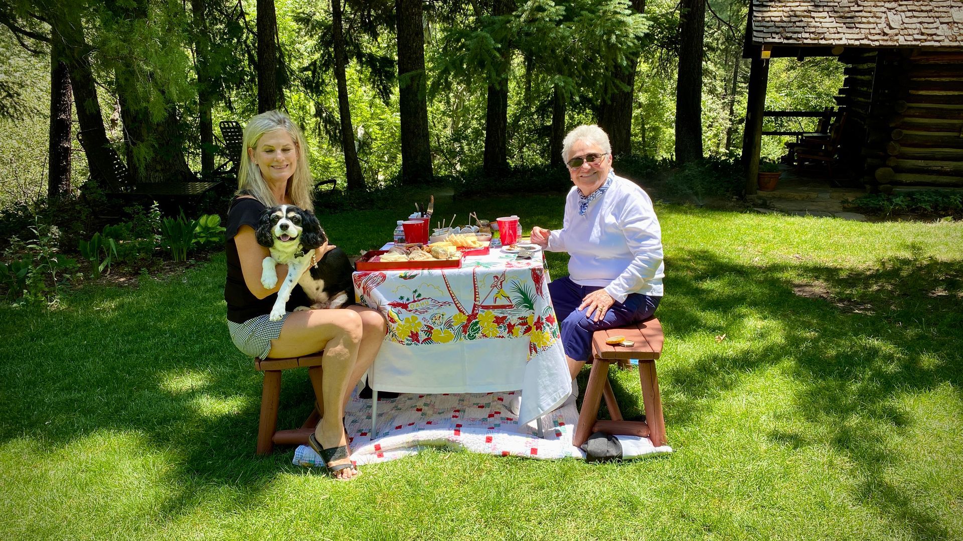 Two people seated at a small table in a sunny garden, with a tea set and flowers on the table.