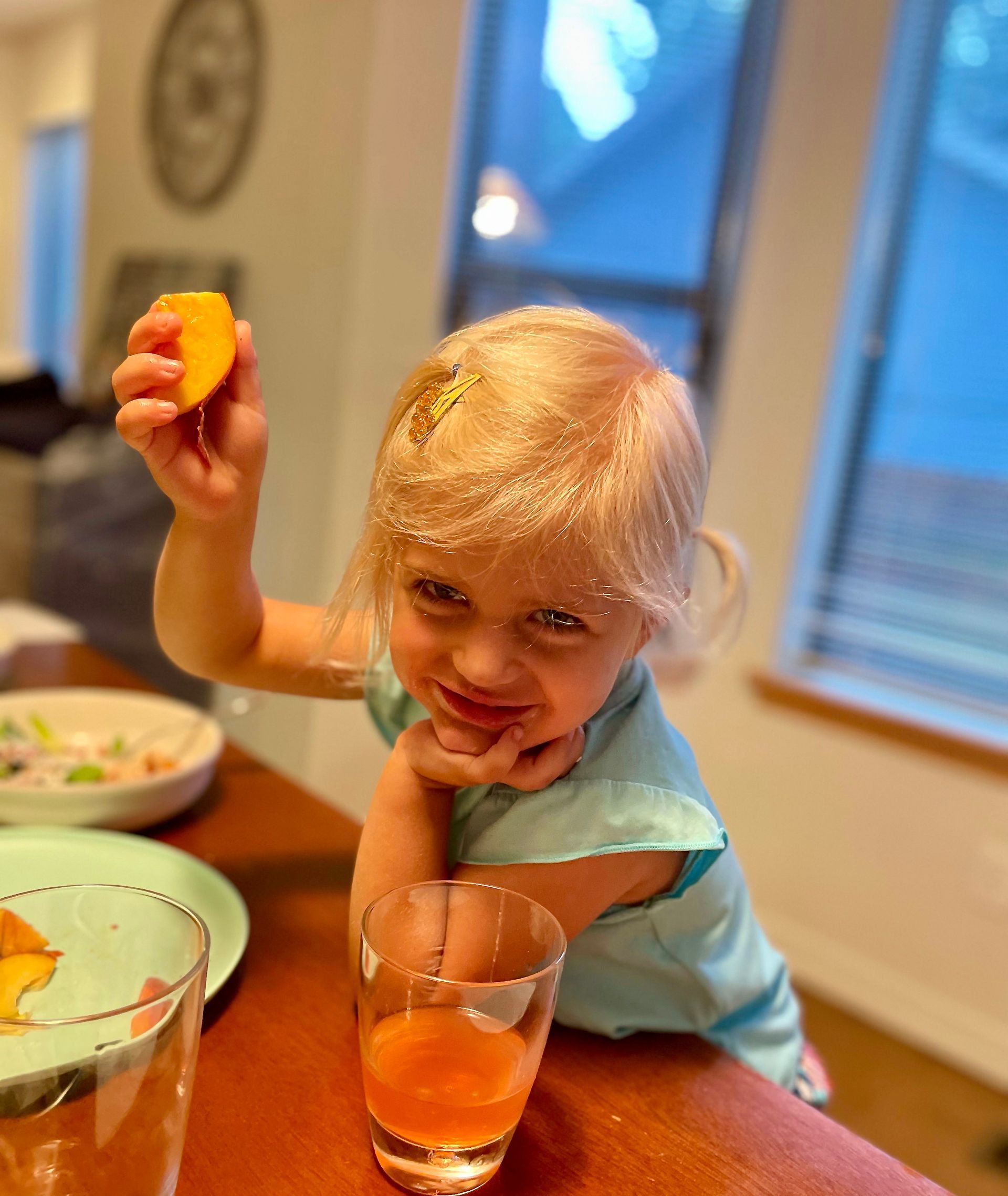 Smiling child at a table holding a fruit slice and a glass of orange drink in a kitchen