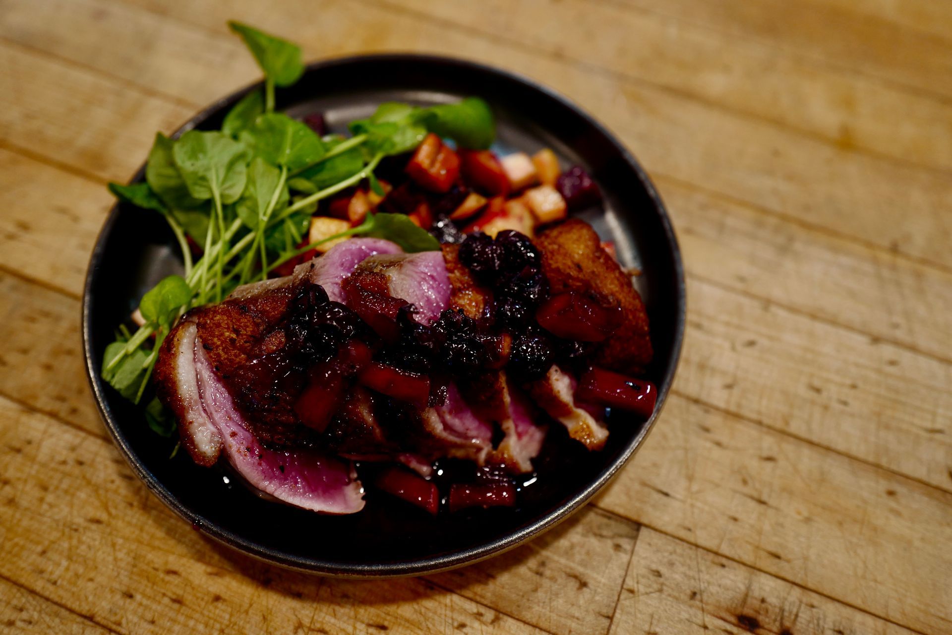 Bowl of glazed meat with vegetables on a wooden table, topped with fresh greens