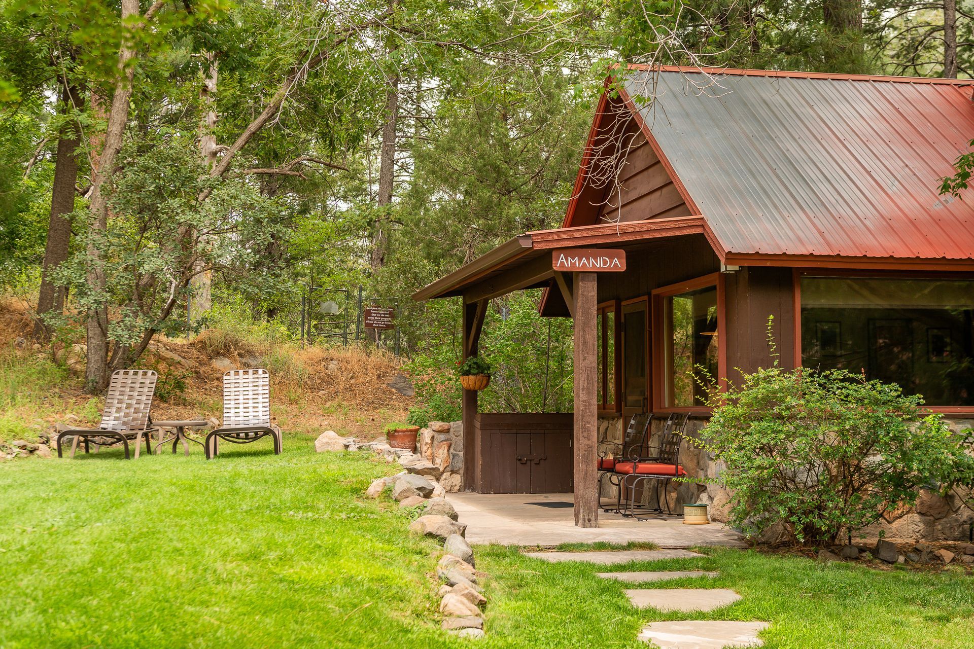 Small rustic cabin with red roof beside a green lawn and benches in a wooded park