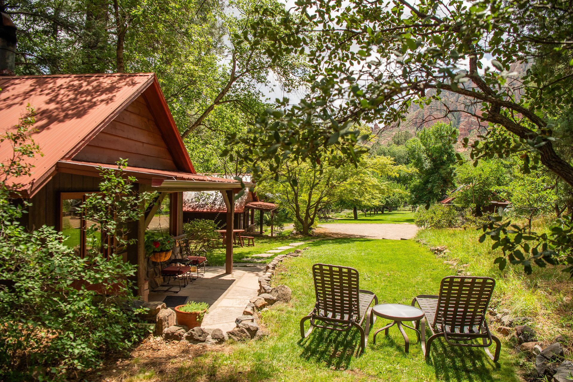 Rustic cabin garden with two chairs on a sunny lawn surrounded by trees and flowers