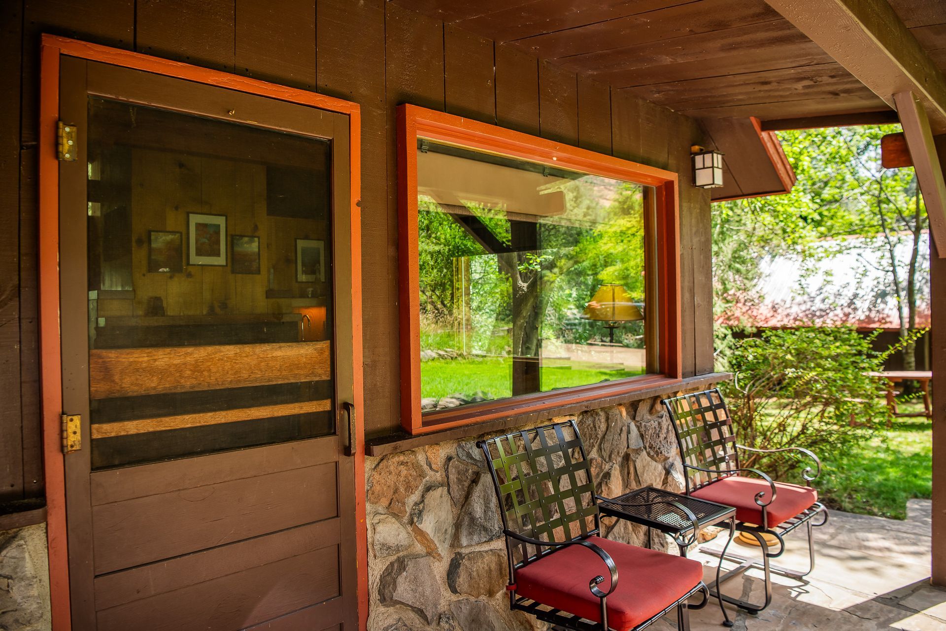 Covered porch with stone wall, glass door, red chairs, and a view of a green garden outdoors