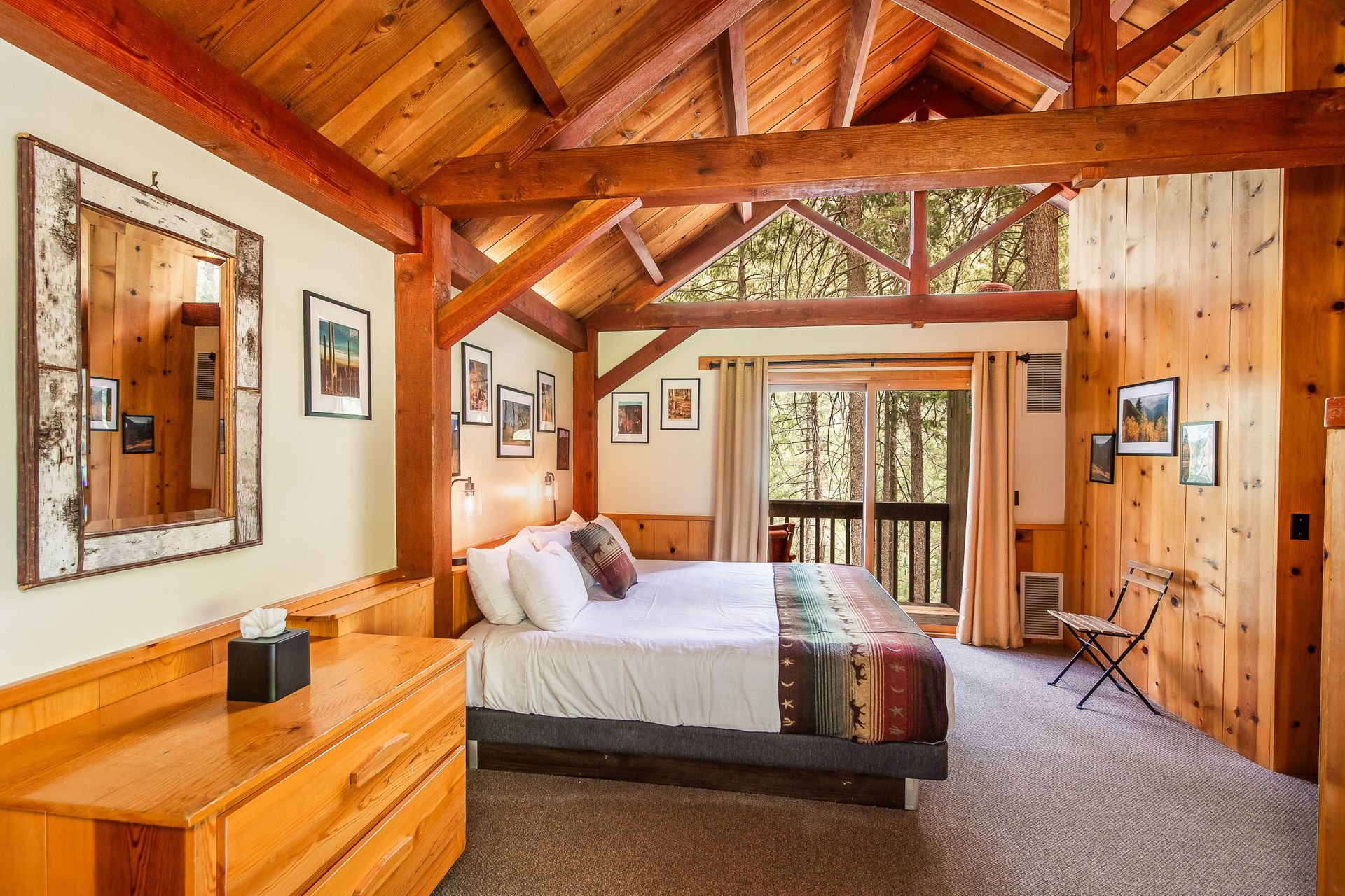 Wood-paneled bedroom with vaulted ceiling, bed, dresser, and large windows overlooking trees