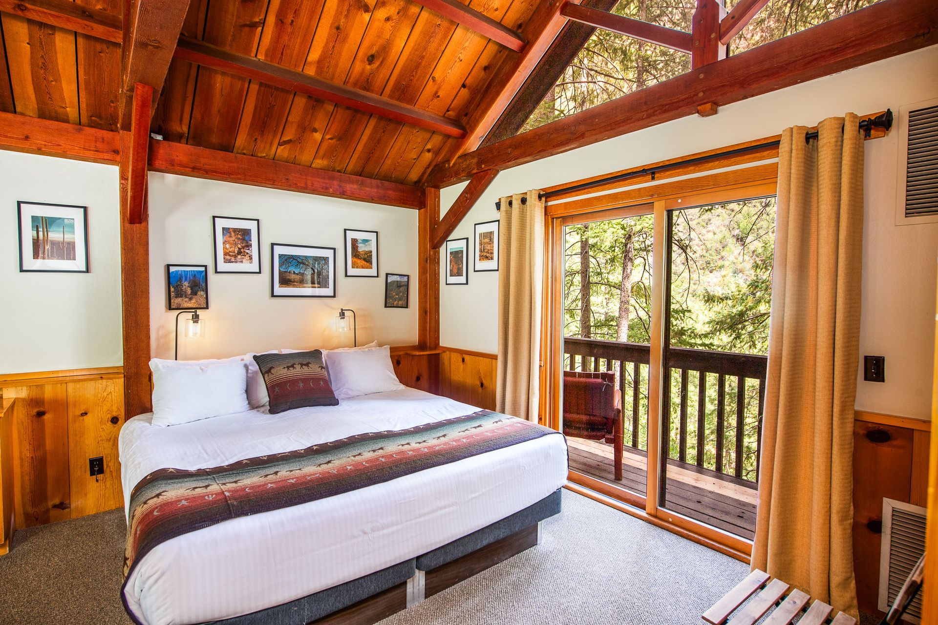 Cozy bedroom with wooden ceiling, white bed, framed photos, and sliding door to a balcony and forest view