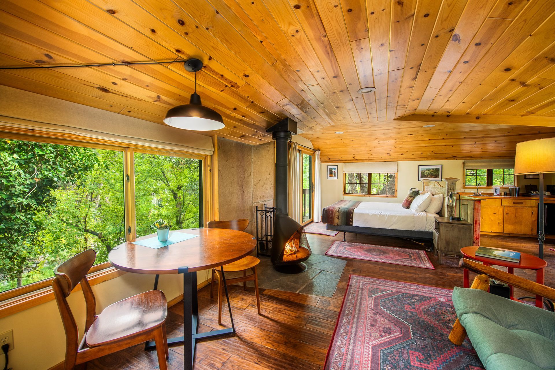 Cozy cabin interior with wood-paneled ceiling, wood stove, bed, dining table, and large window views of greenery