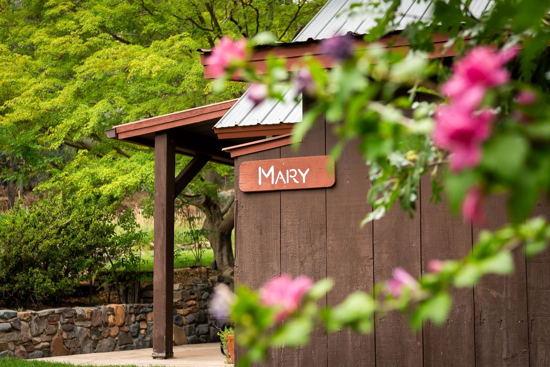 Rustic sign reading “MAY” on a wooden wall, with pink flowers and green trees in the background