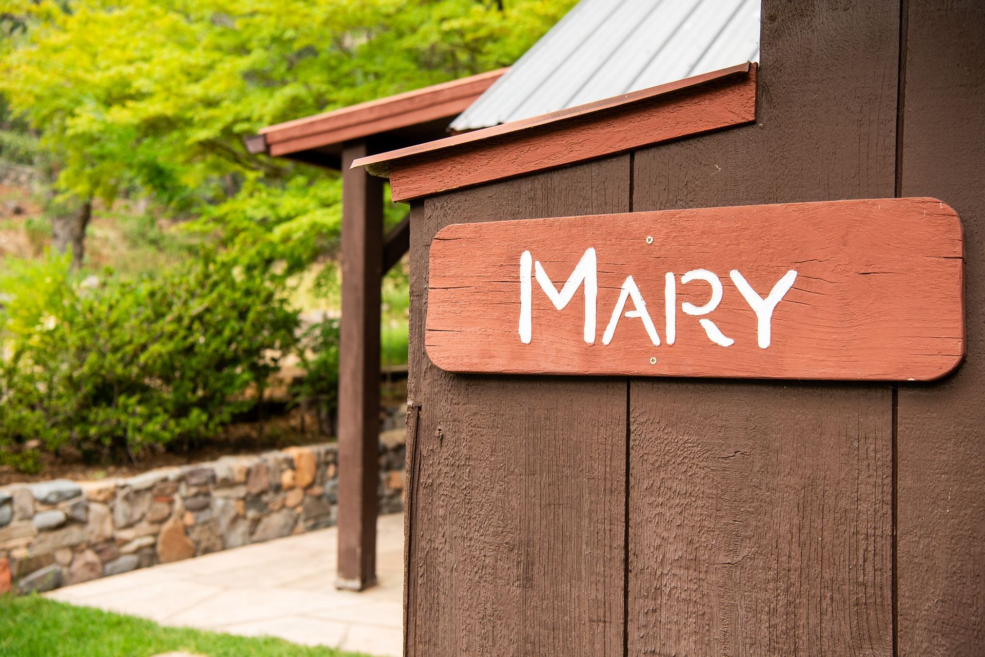 Rustic sign reading “MARY” on a brown building beside a path and greenery