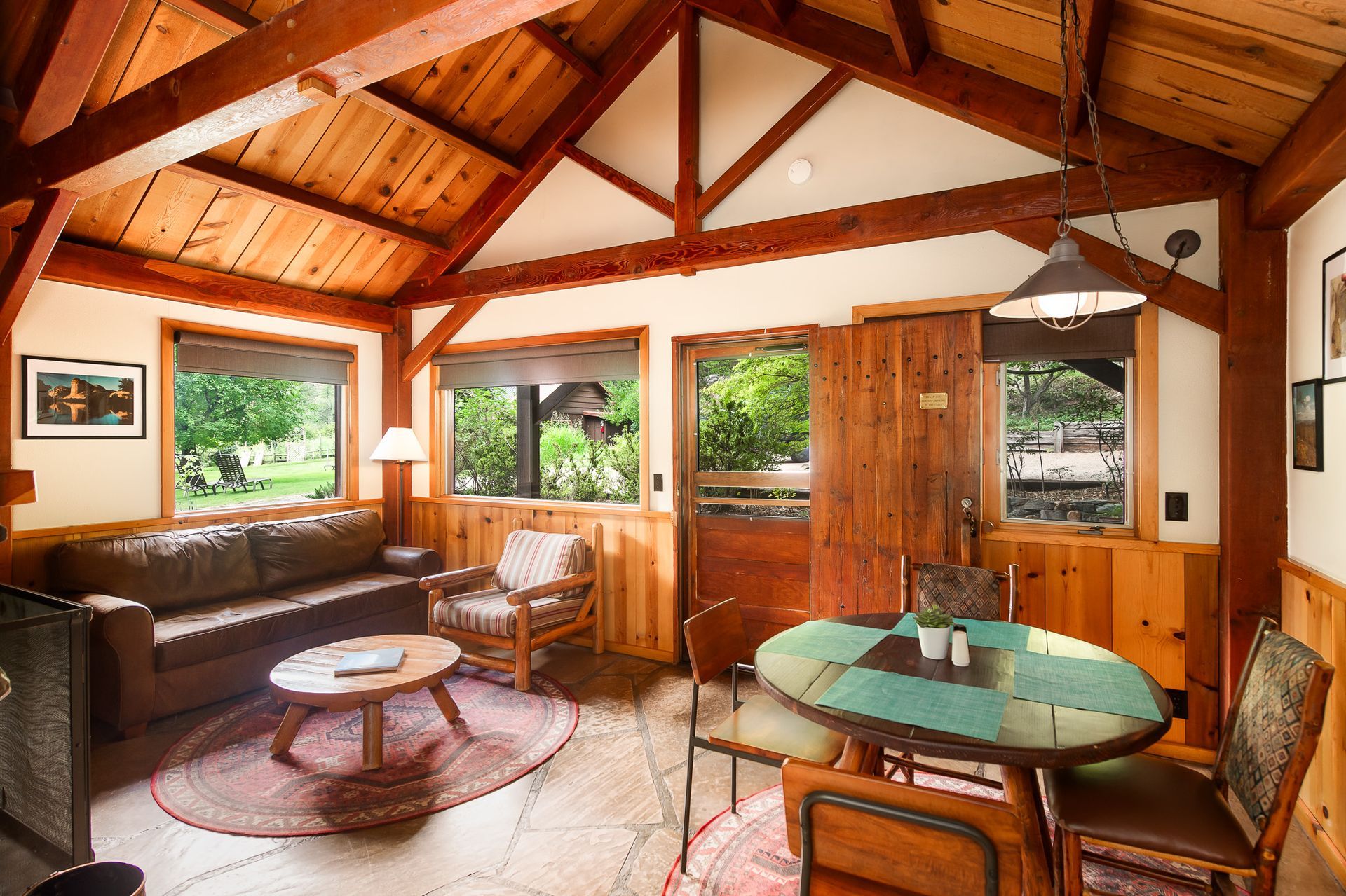 Cozy wooden cabin living room with sofa, chairs, round table, and large windows overlooking greenery