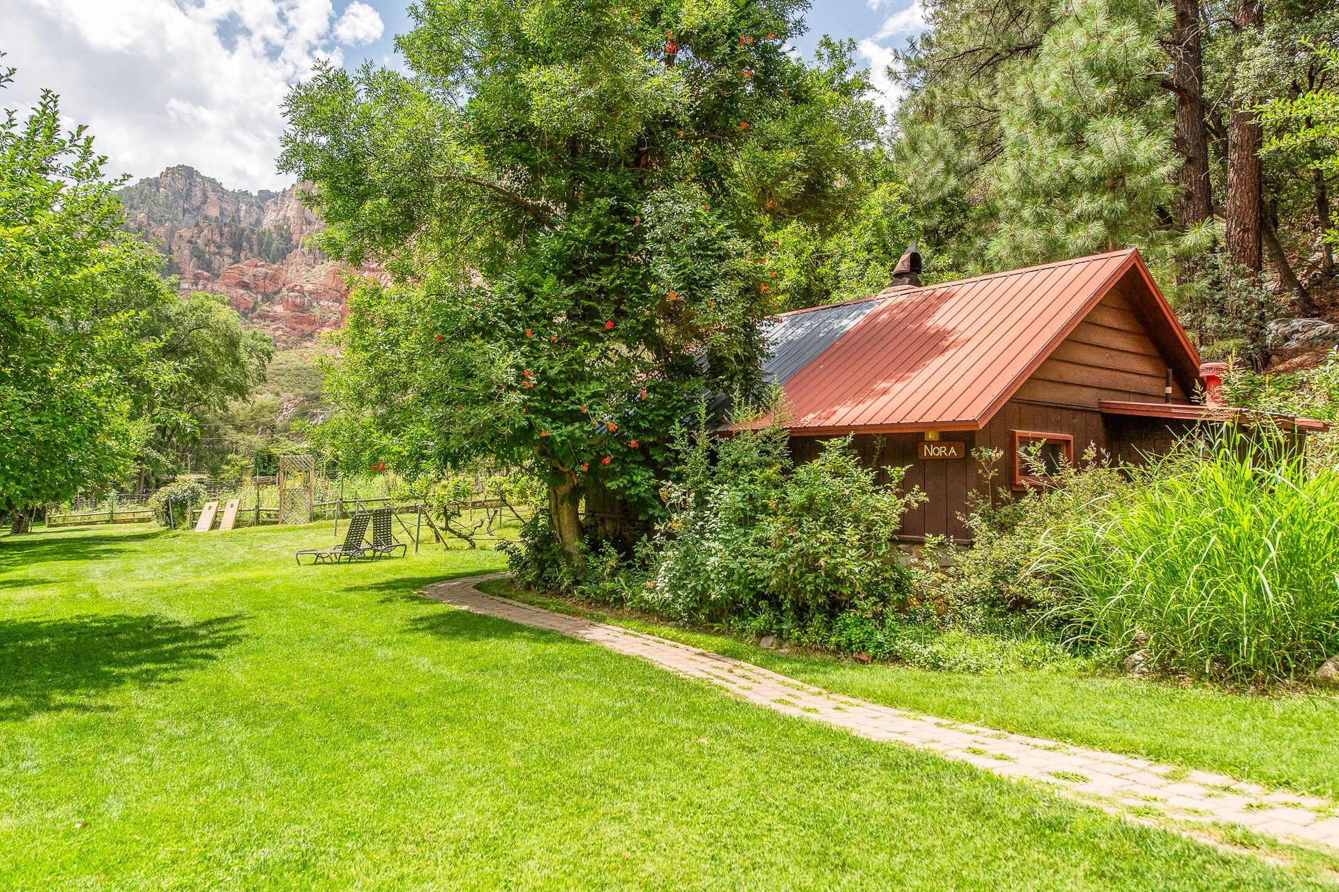 A small wooden cabin with a red roof beside a grassy path in a sunny green forest clearing.