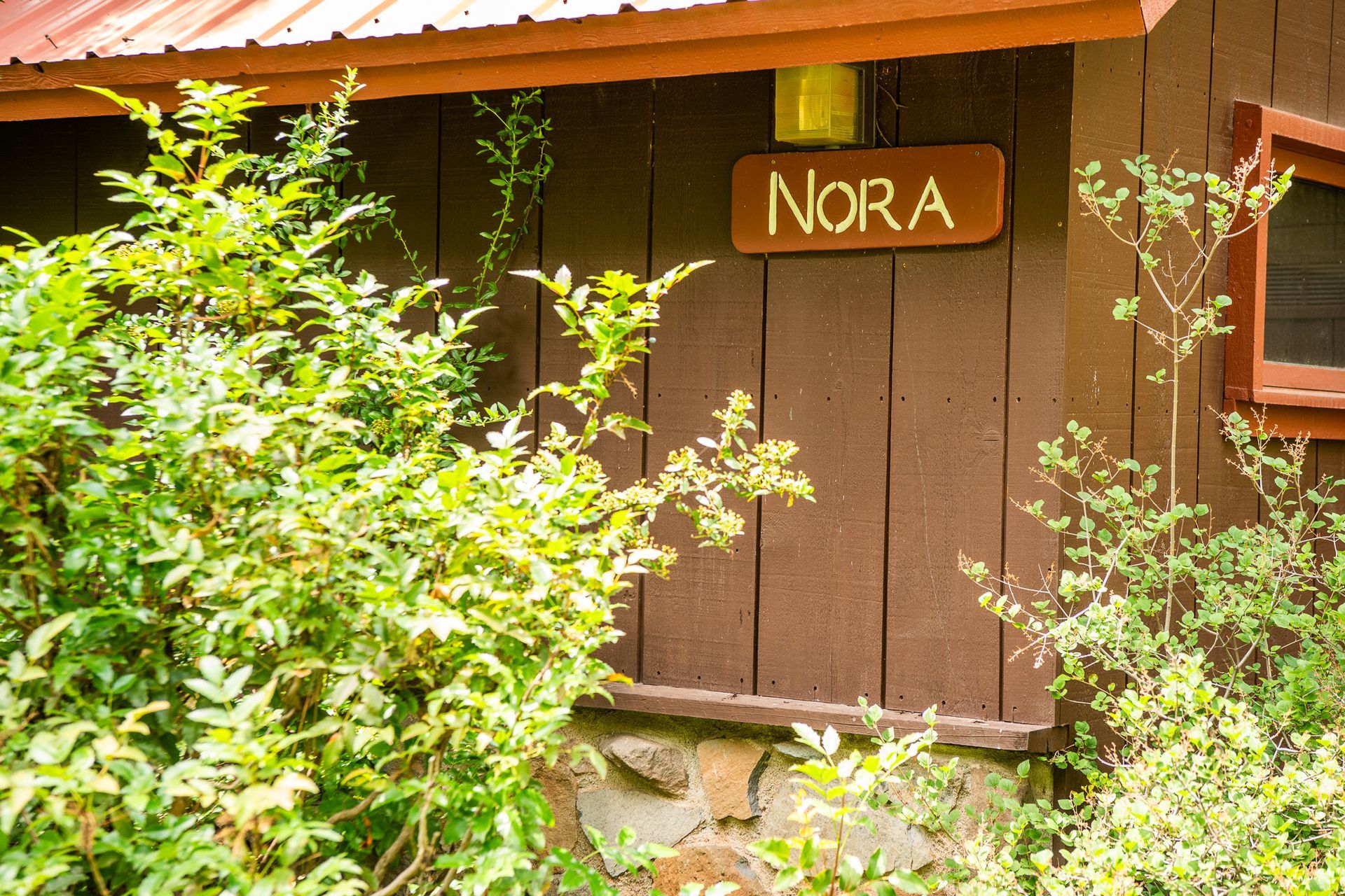 Wooden cabin wall with a sign reading “Nora,” partly hidden by green bushes.