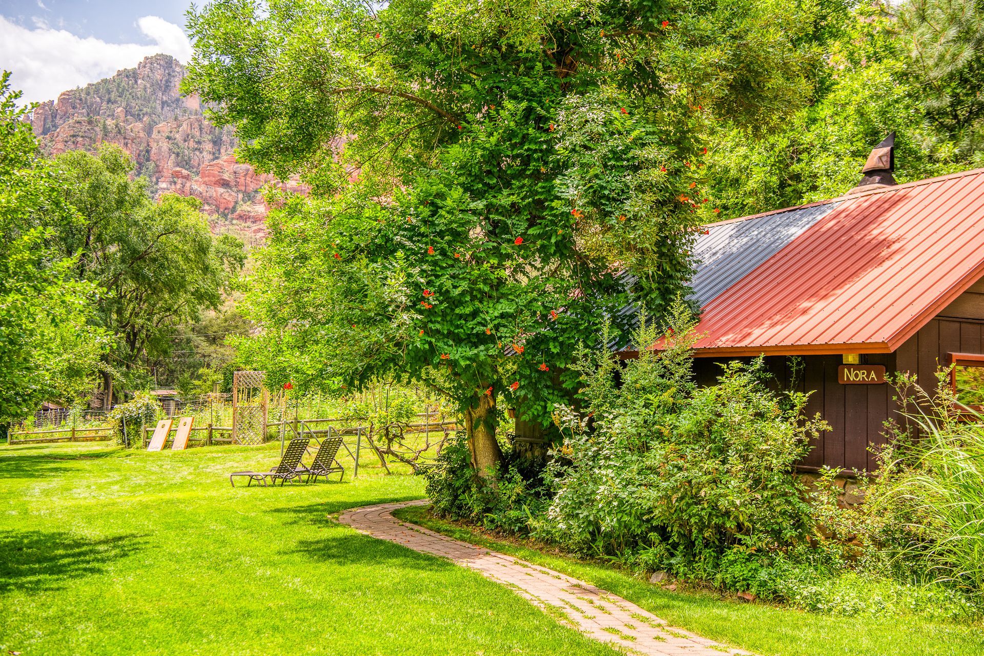 Sunny garden with green lawn, trees, and a small house with a red roof by a mountain backdrop