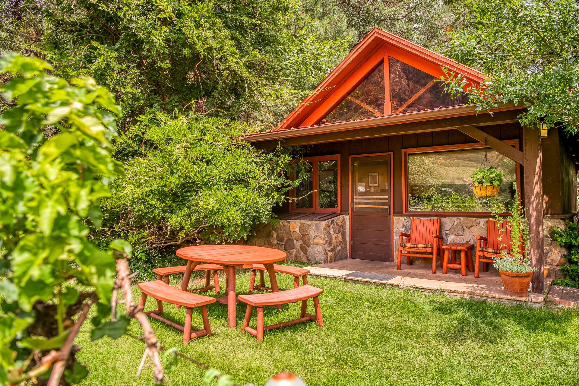 Small wooden cabin with a porch and red outdoor table and chairs in a lush green garden