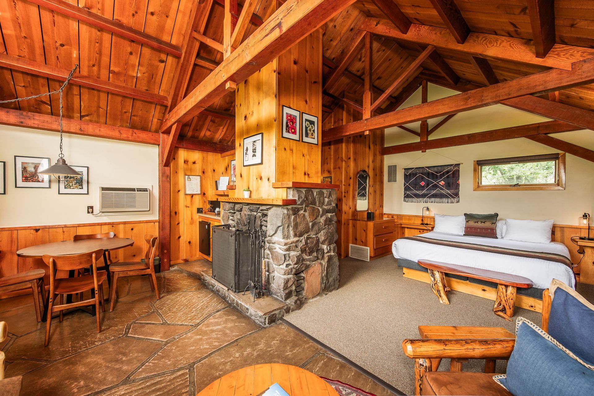 Rustic wooden cabin interior with stone fireplace, dining table, and a bed by the windows