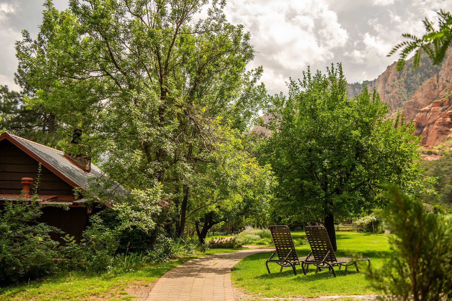 Sunny garden path beside a wooden cabin, with trees, lawn chairs, and mountains in the background