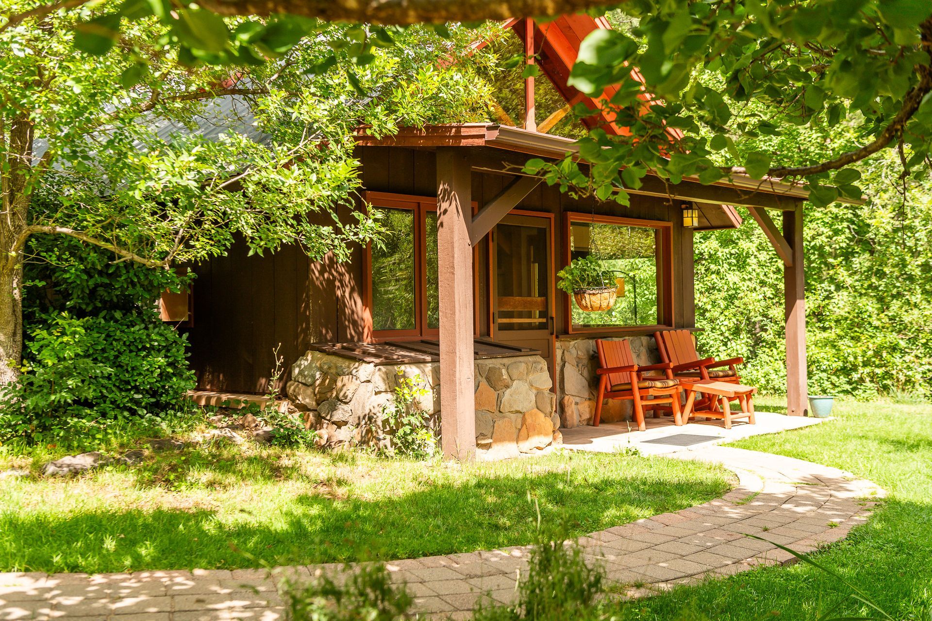 Wooden cottage porch nestled in lush green garden, with stone path and patio chairs under trees.