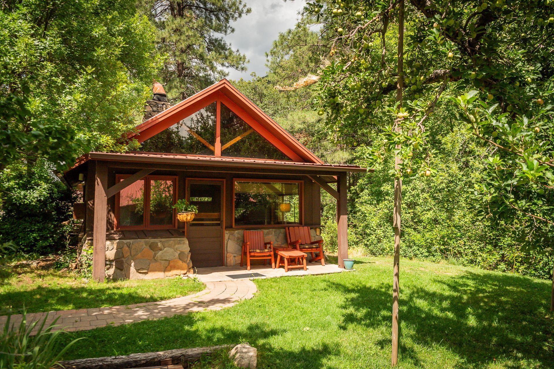 Small wooden cabin with a red roof in a sunny green garden, surrounded by trees.