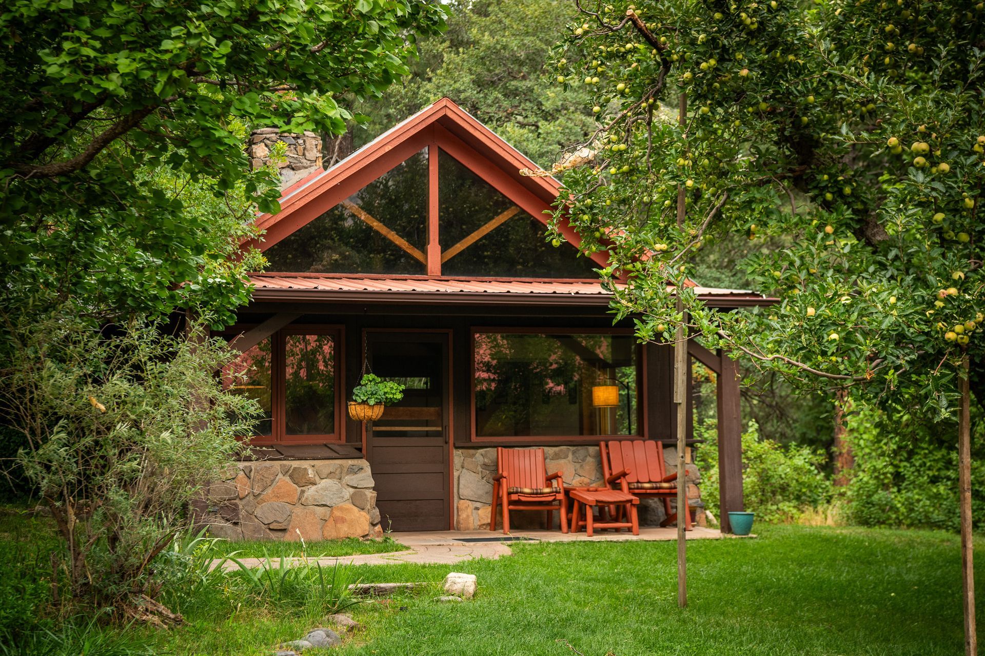 Small rustic cabin with a porch and red chairs, surrounded by lush green trees and grass.