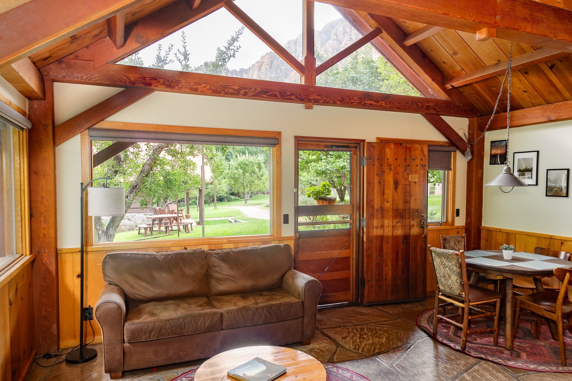 Wooden cabin living room with sofa, dining table, and large window overlooking a green yard