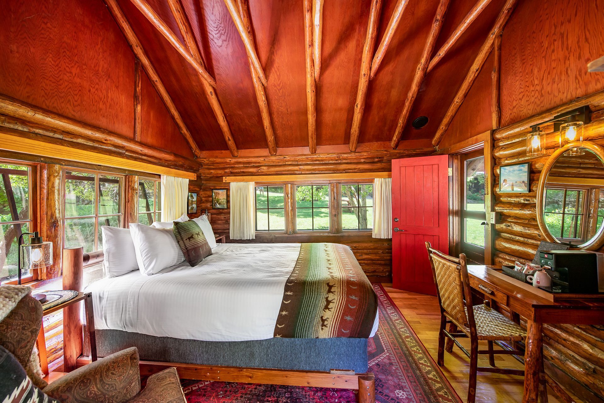 Rustic bedroom with a large bed, wood-beam ceiling, and desk by windows overlooking greenery