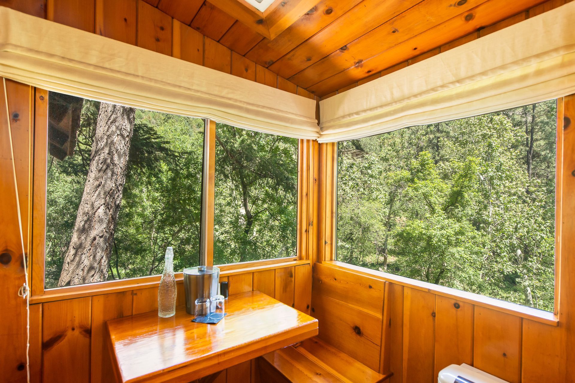 Wood-paneled sunroom with large windows, a corner bench, and a wooden table with bottles.
