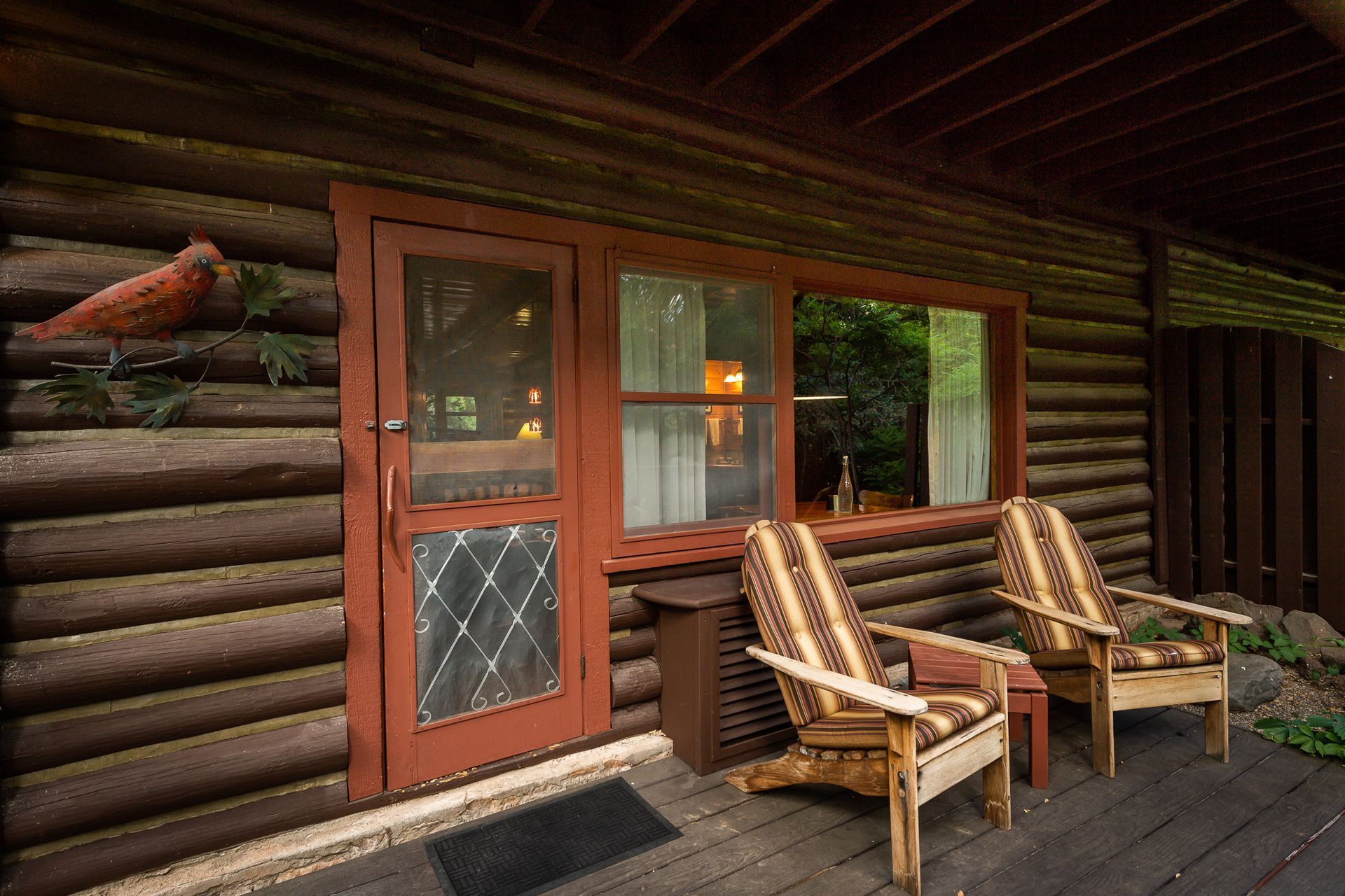 Rustic cabin porch with two wooden chairs and a small table beside a log wall and window.