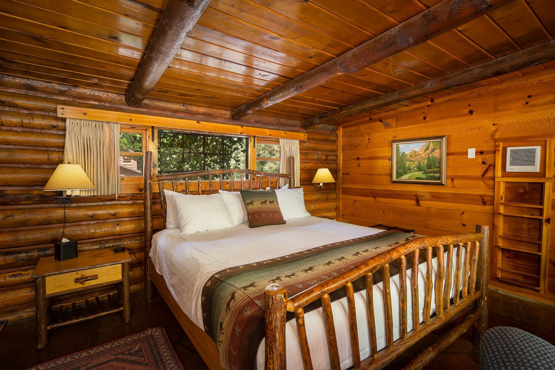 Rustic log cabin bedroom with wood-paneled walls, a bed, bedside lamps, and a window view of trees