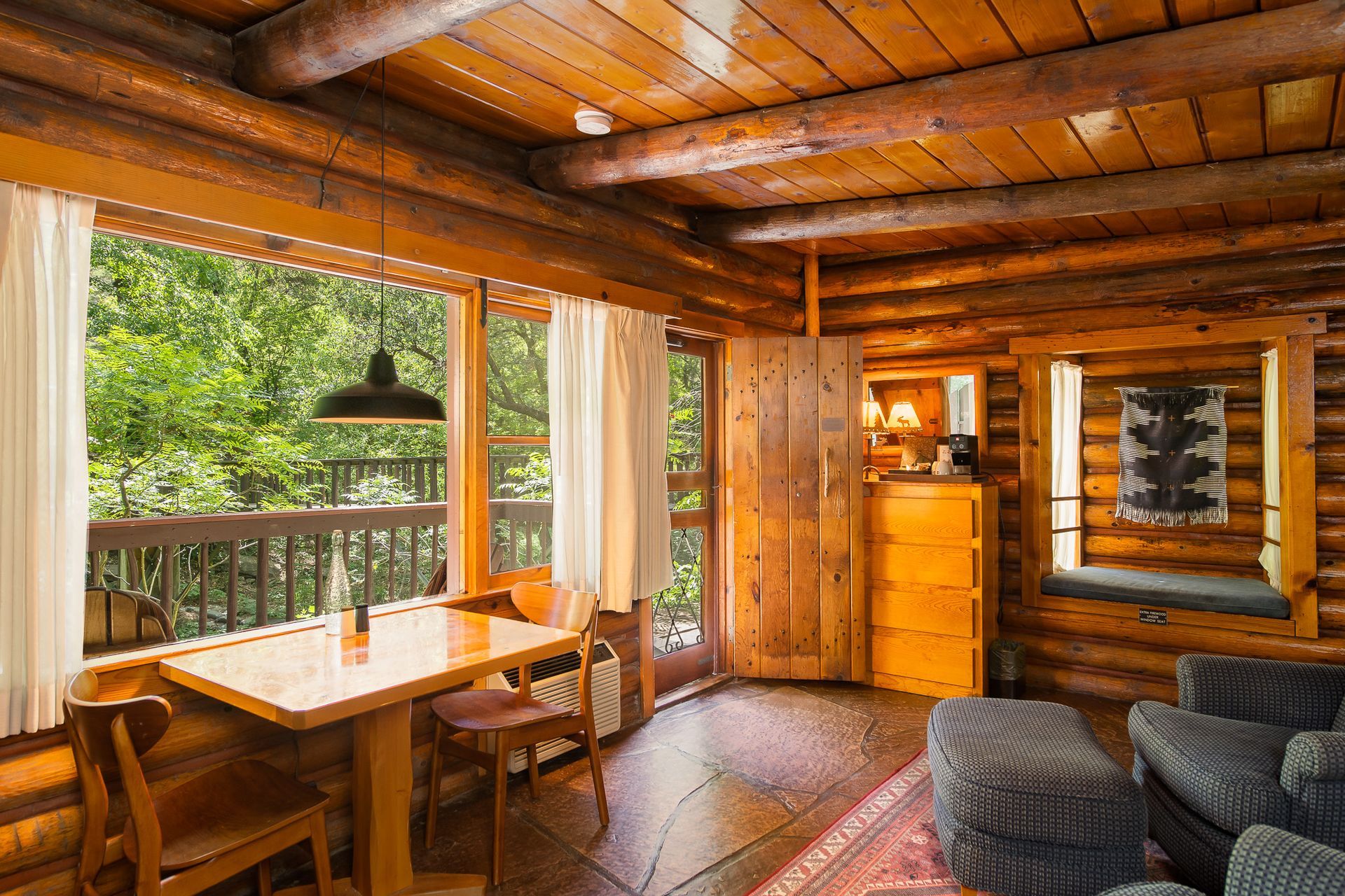 Rustic wooden cabin interior with dining table, chairs, sofa, and balcony view through large windows