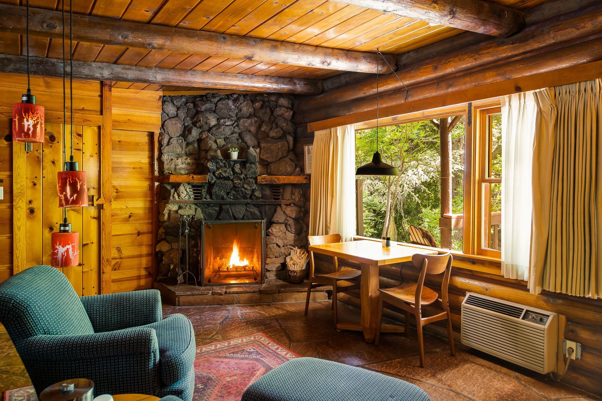 Cozy log cabin living room with stone fireplace, wood-paneled walls, and large windows overlooking trees.