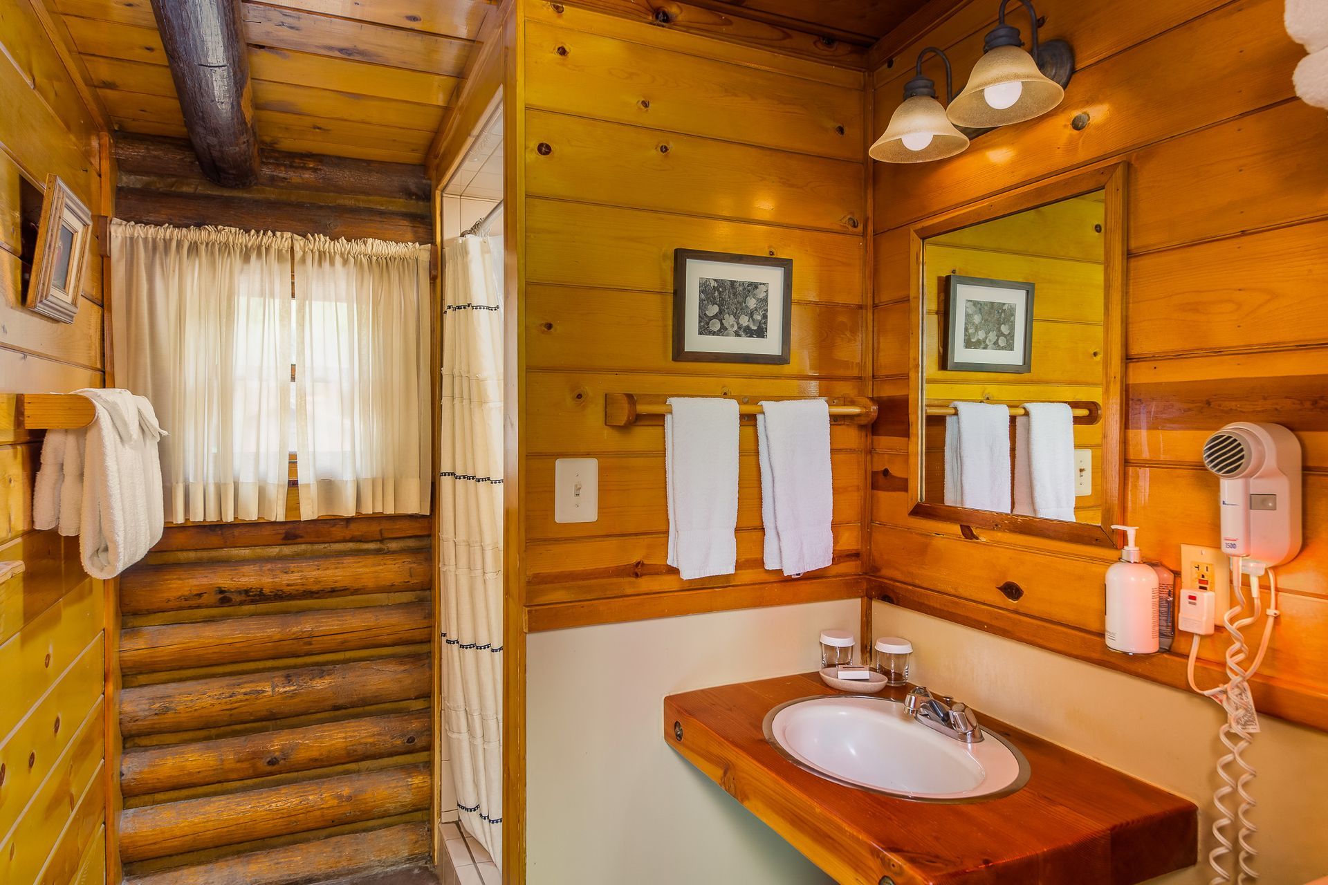 Wood-paneled bathroom with sink, mirror, towels, and a stairway beside a curtained window.