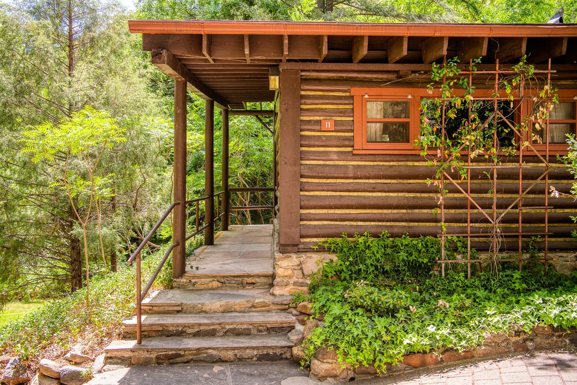 Wooden cabin porch with stone steps, a small window, and greenery beside a forested path