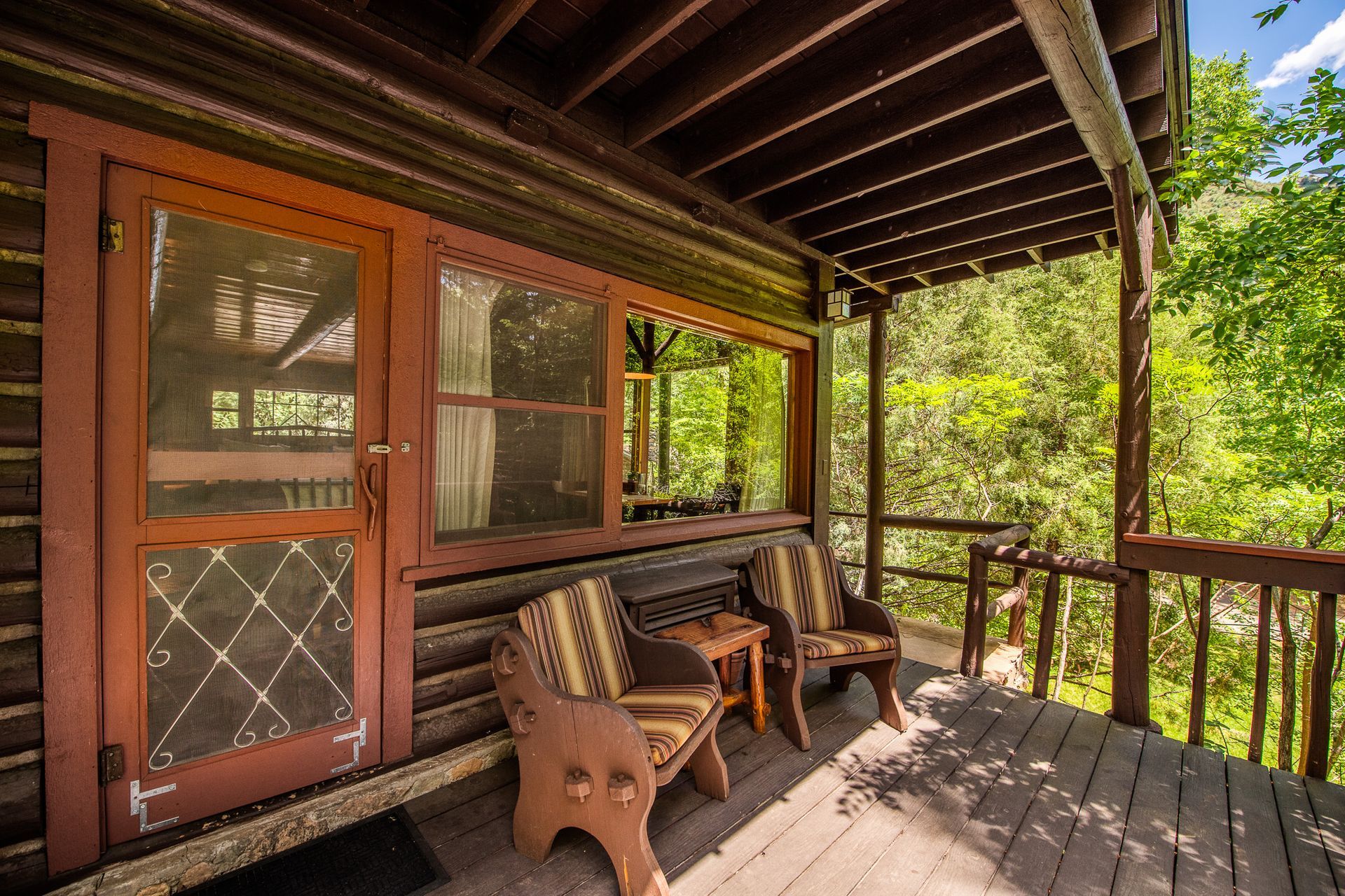 Wooden porch with bench and chairs beside a screened door, overlooking a green wooded area.