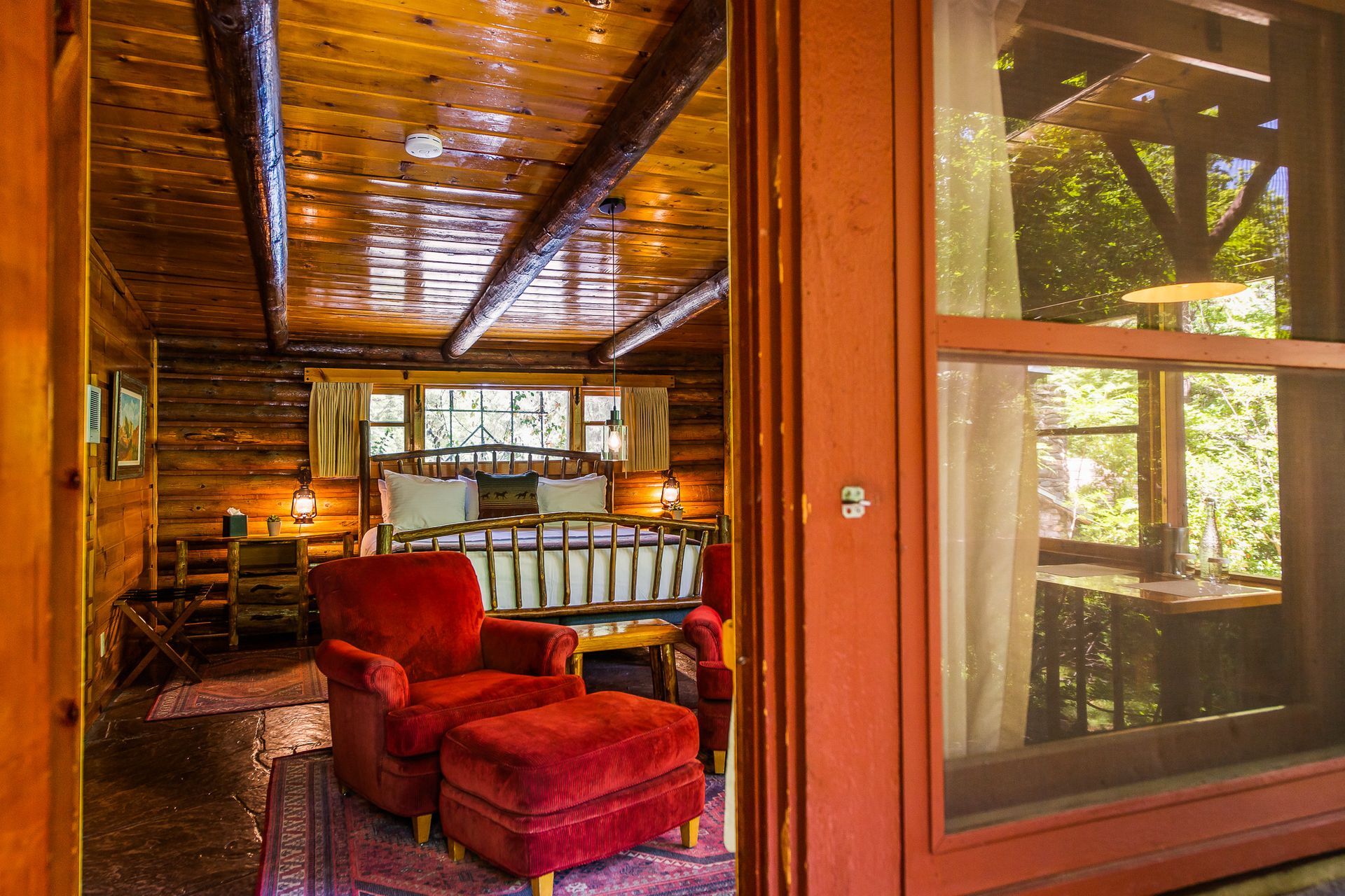 Wood-paneled cabin interior with red chair and ottoman, viewed through an open door to a forest deck.