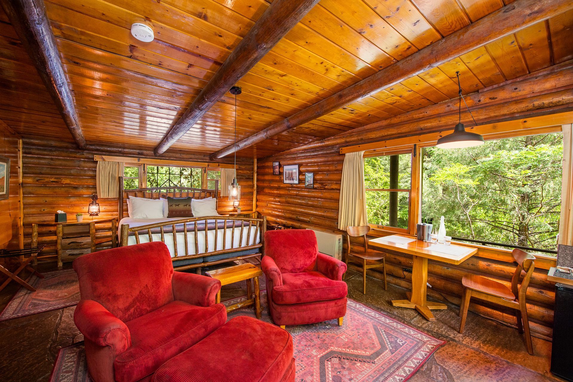 Cozy log cabin living room with red armchairs, wooden ceiling, and large windows overlooking greenery