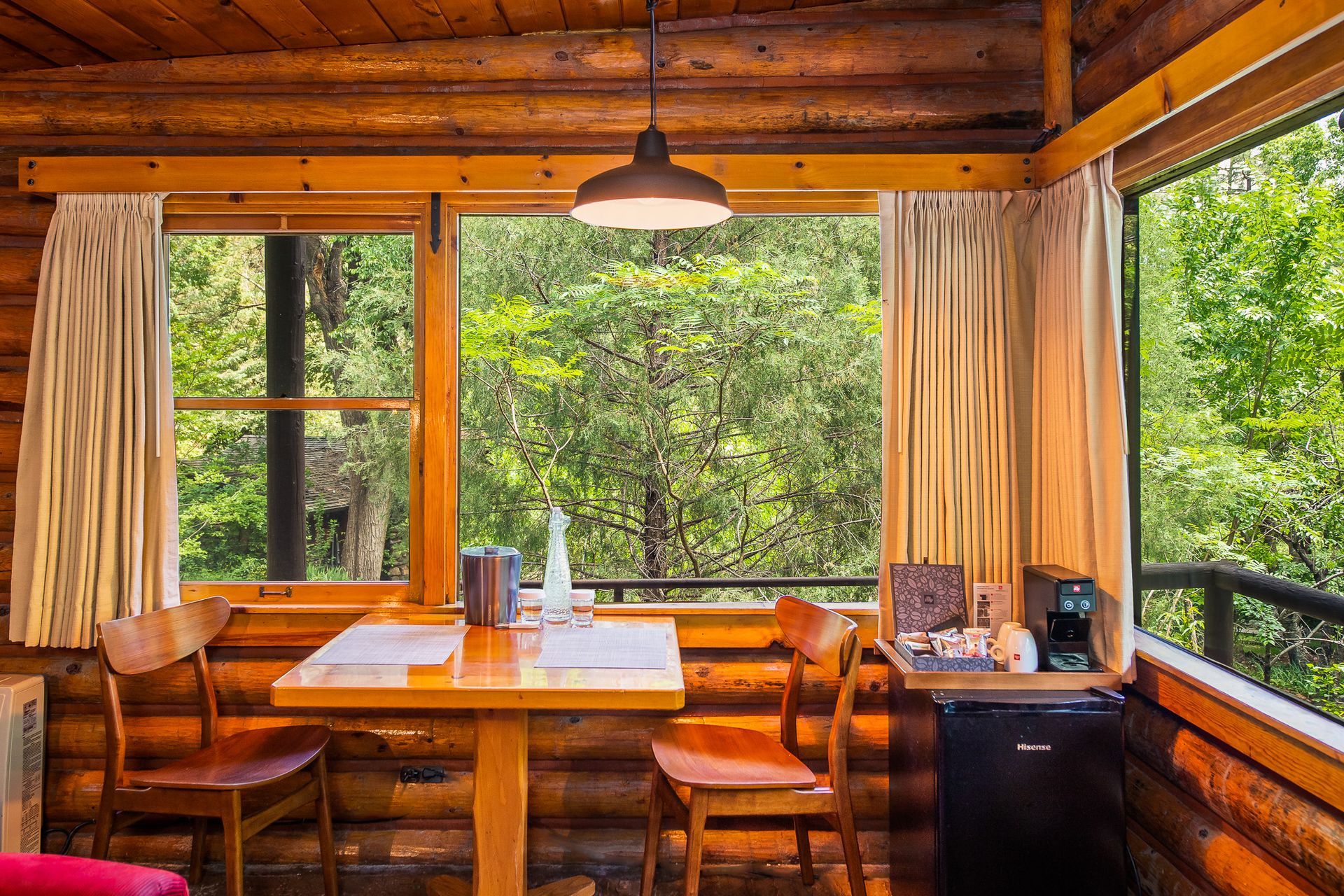 Rustic cabin dining nook with wooden table, chairs, and open windows overlooking green trees