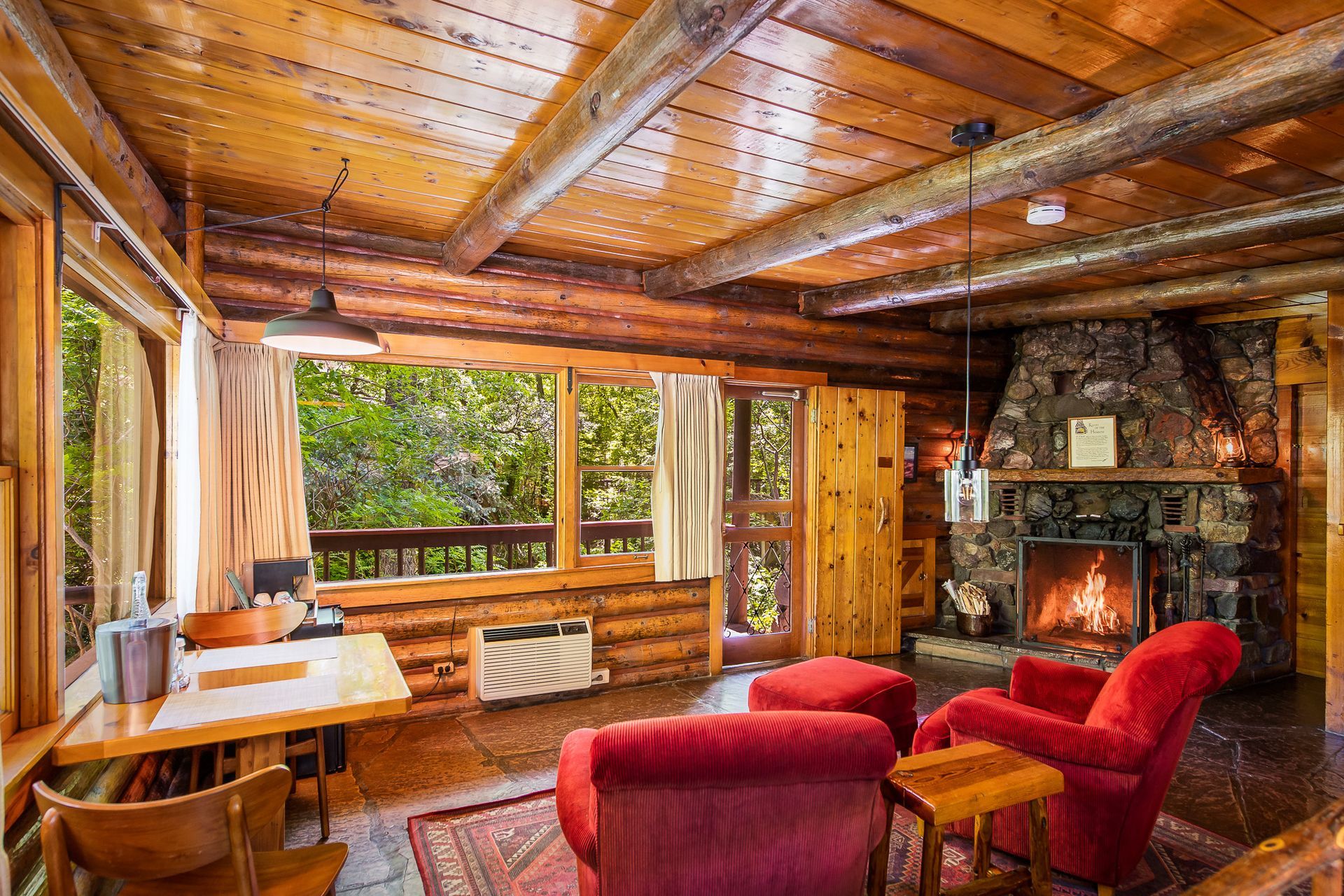 Cozy cabin living room with red chairs, wood-paneled ceiling, and a lit fireplace overlooking greenery