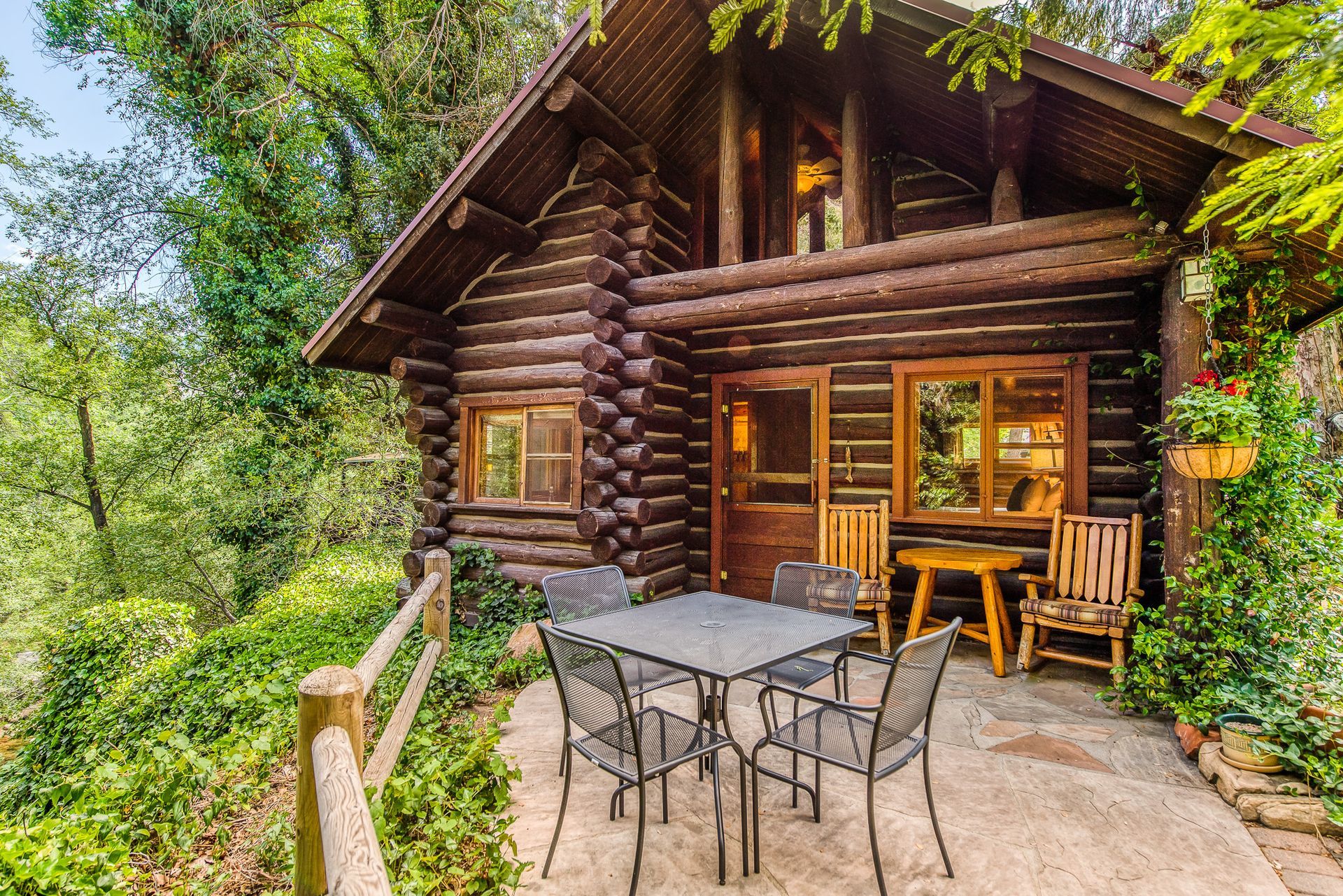 Log cabin patio with outdoor dining table and chairs, surrounded by lush green trees.