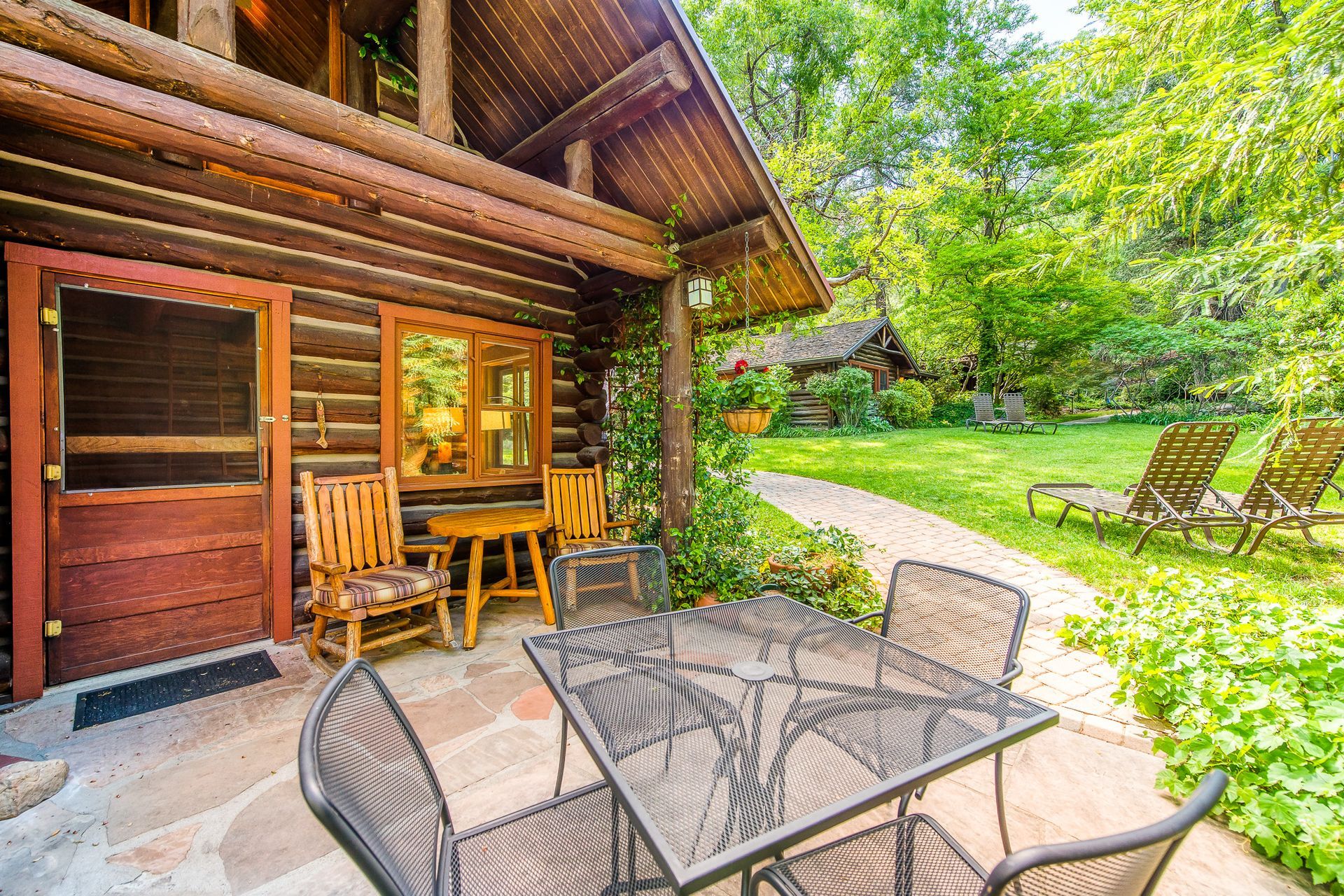 Wooden cabin patio with metal table and chairs, overlooking a sunny green garden with a bench.