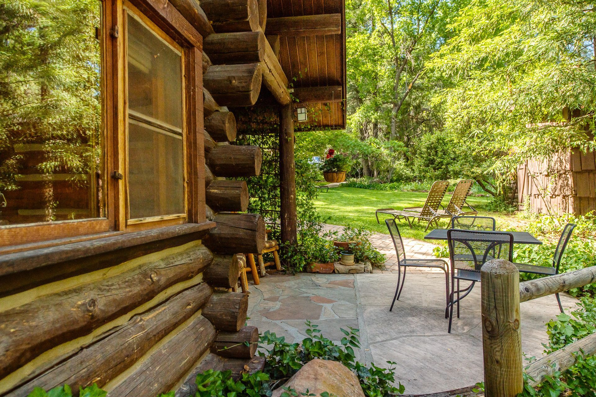 Log cabin patio with metal table and chairs amid green trees and stone path