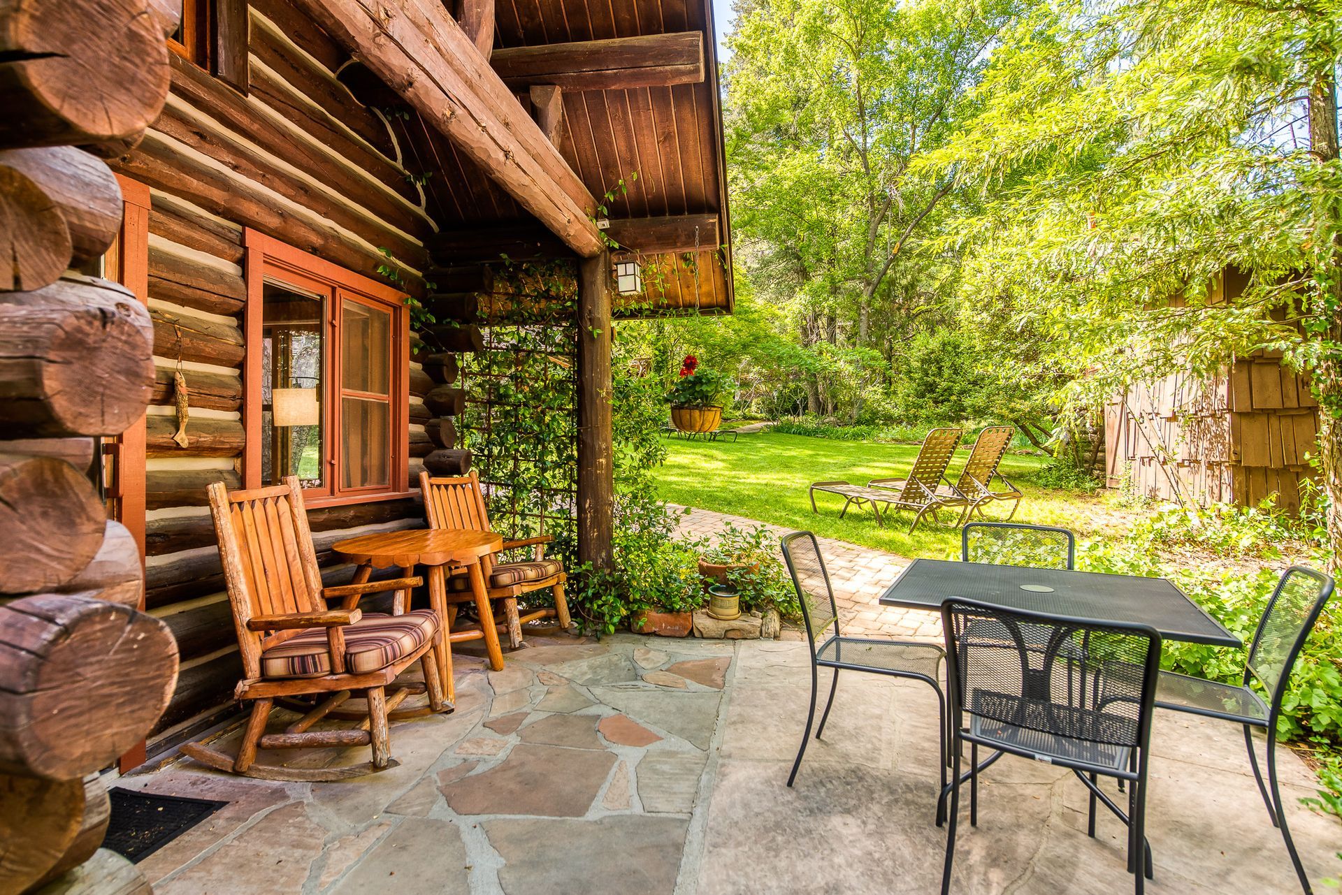Rustic log cabin patio with wooden chairs and metal table beside a green garden