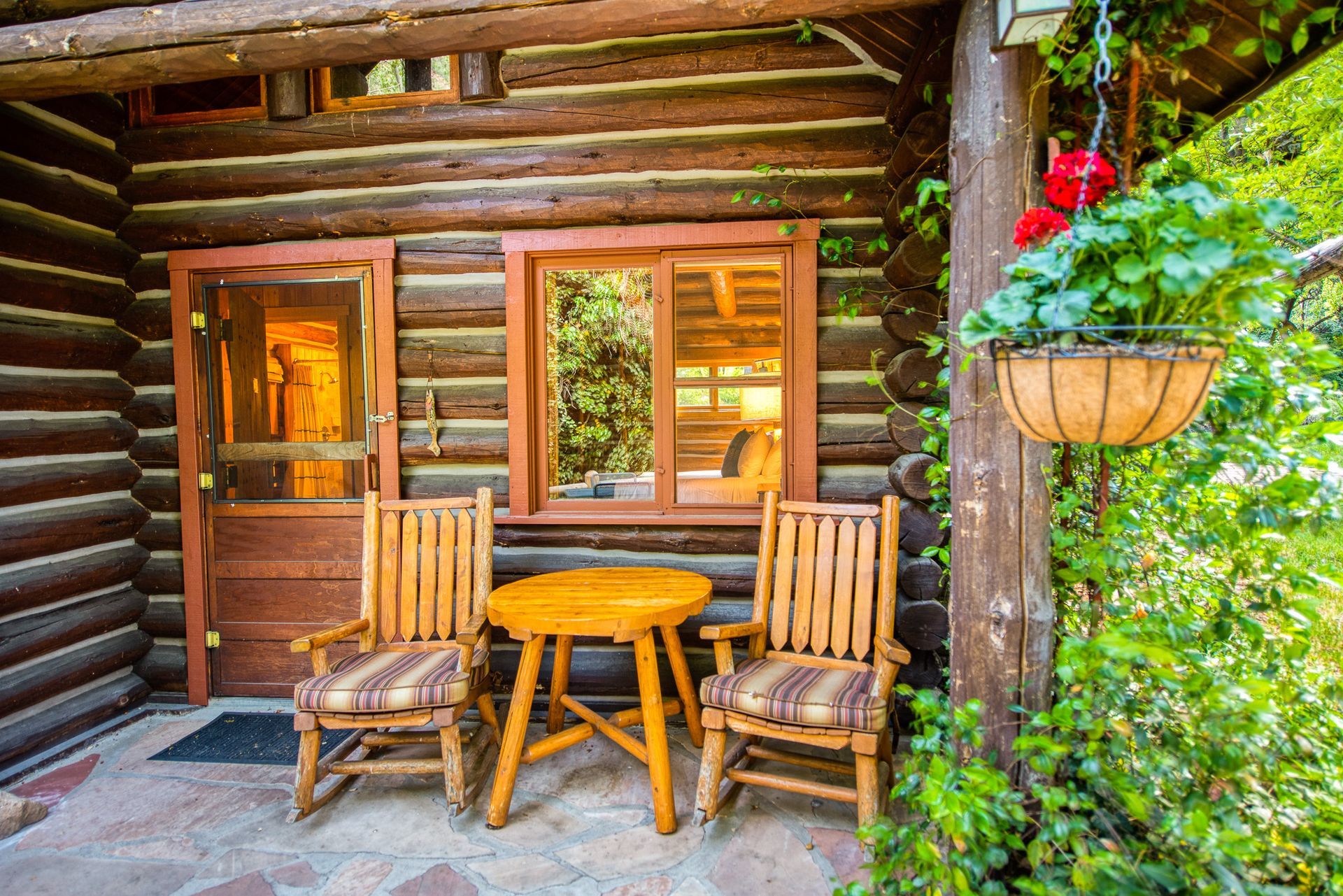 Rustic cabin porch with two wooden chairs, small table, and hanging flower basket beside a log wall