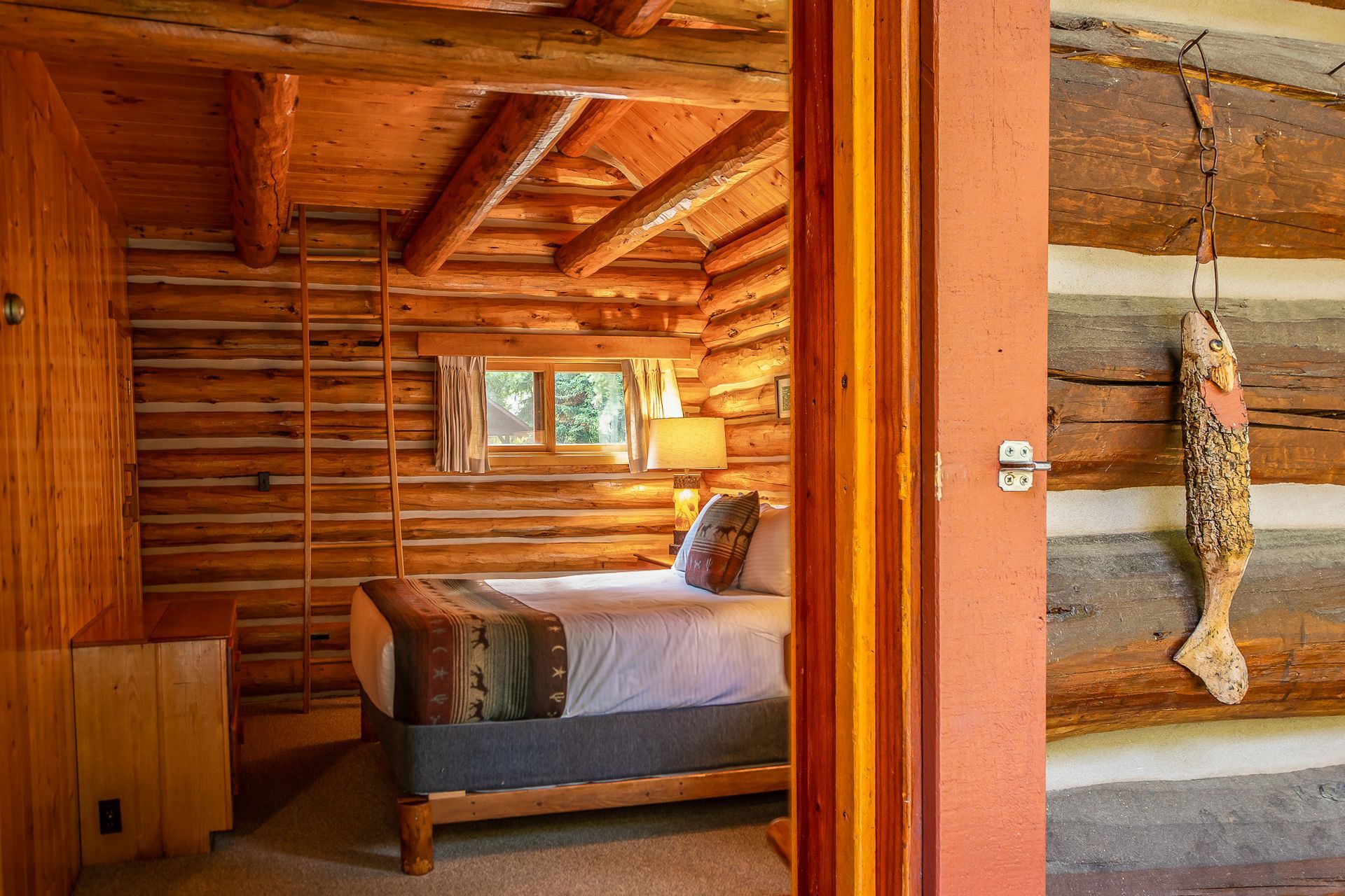 Rustic log cabin bedroom with a bed, window, lamp, and hanging dried fish in the doorway